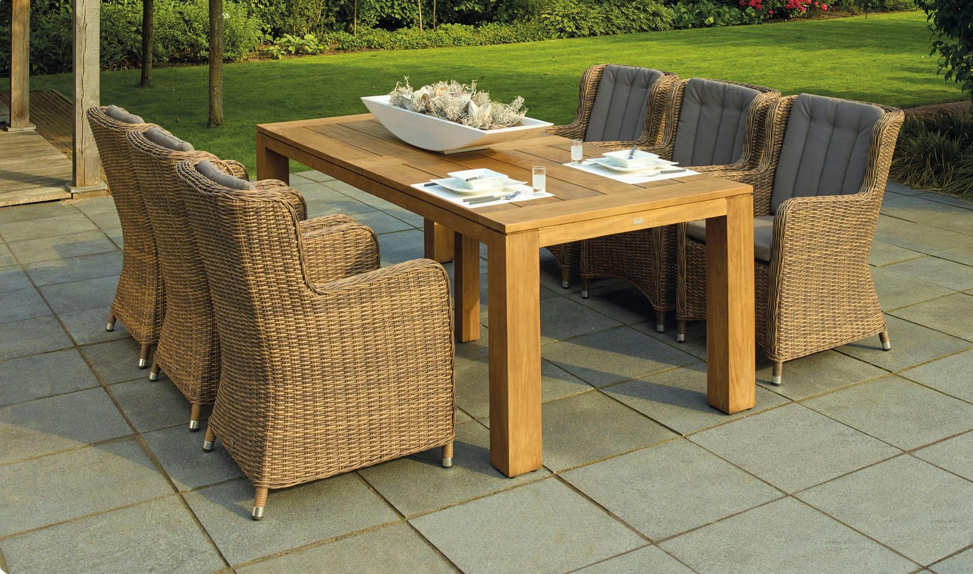 A rectangular wooden patio dining table with six woven wicker chairs featuring grey cushions on a paved terrace.