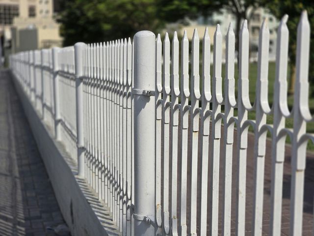 A white metal picket fence stands on a concrete base, stretching into the distance along a path.