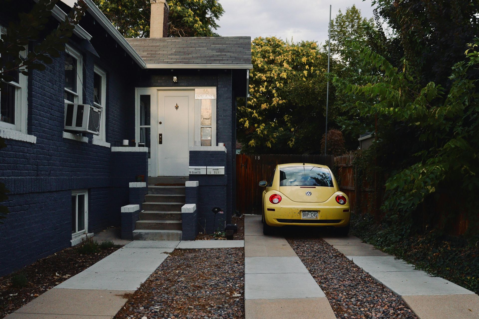 A yellow Volkswagen Beetle parked on a driveway next to a blue, brick house with a white door and small front stairs.