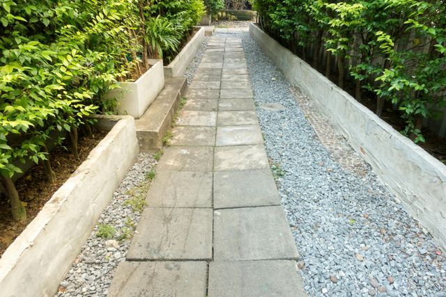 A stone paver walkway bordered by concrete planters, gravel, and green shrubs.