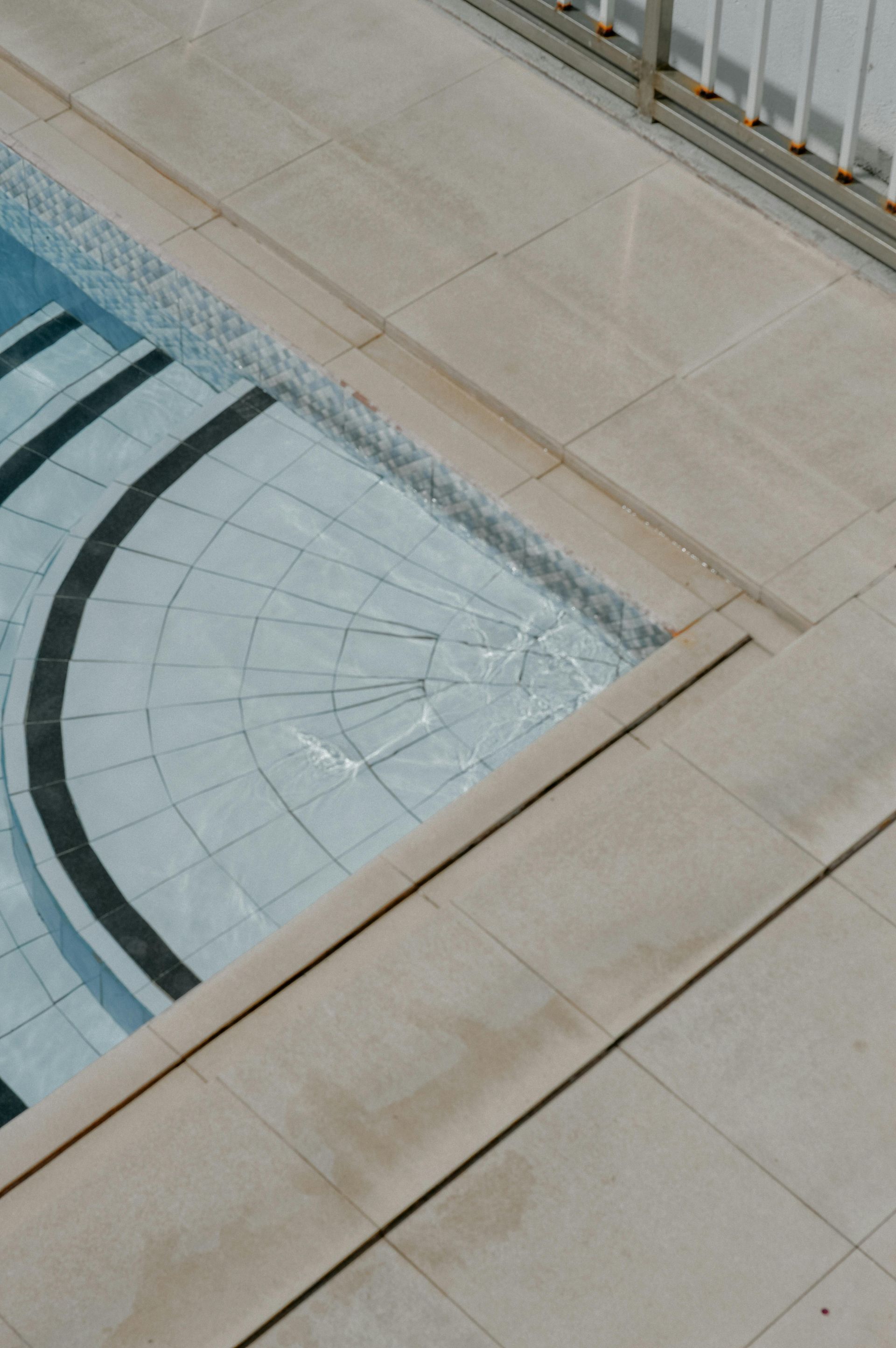 Top-down view of a swimming pool corner with light-colored tile steps, curved dark accents, and pale stone decking.