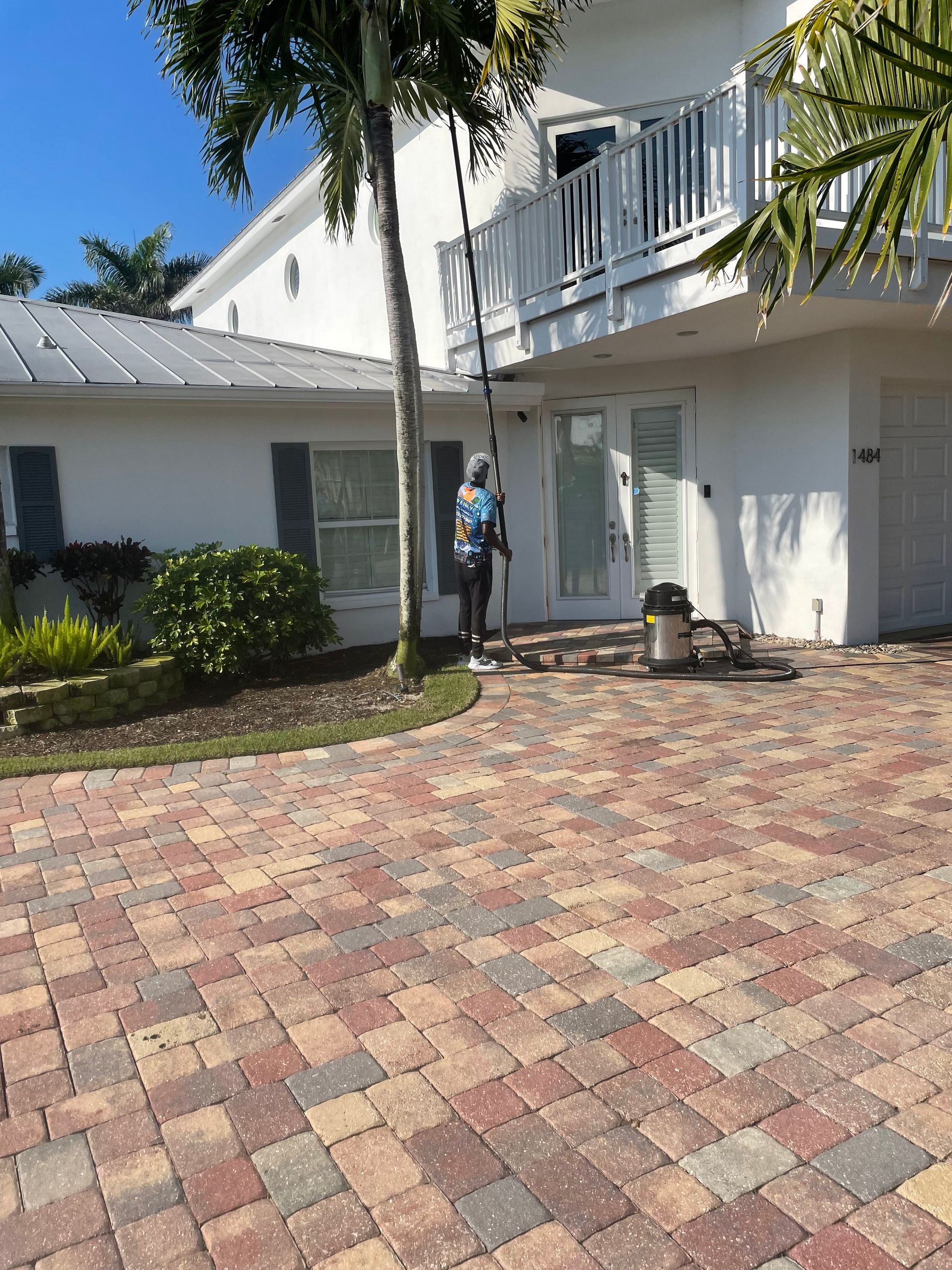 A person uses a tall pole brush to clean the windows of a two-story white house with a paver driveway.