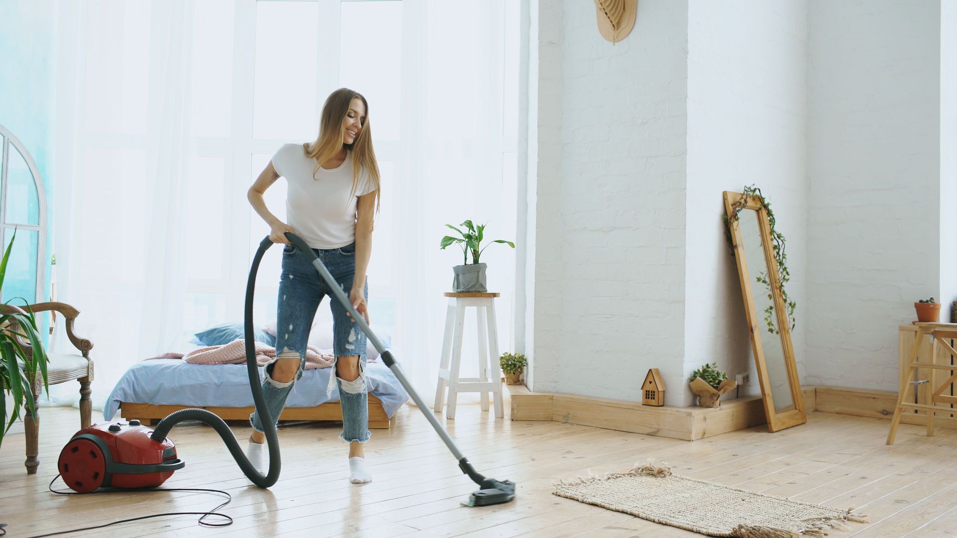 Woman vacuuming a bright living room with a canister vacuum, mirror and plants nearby