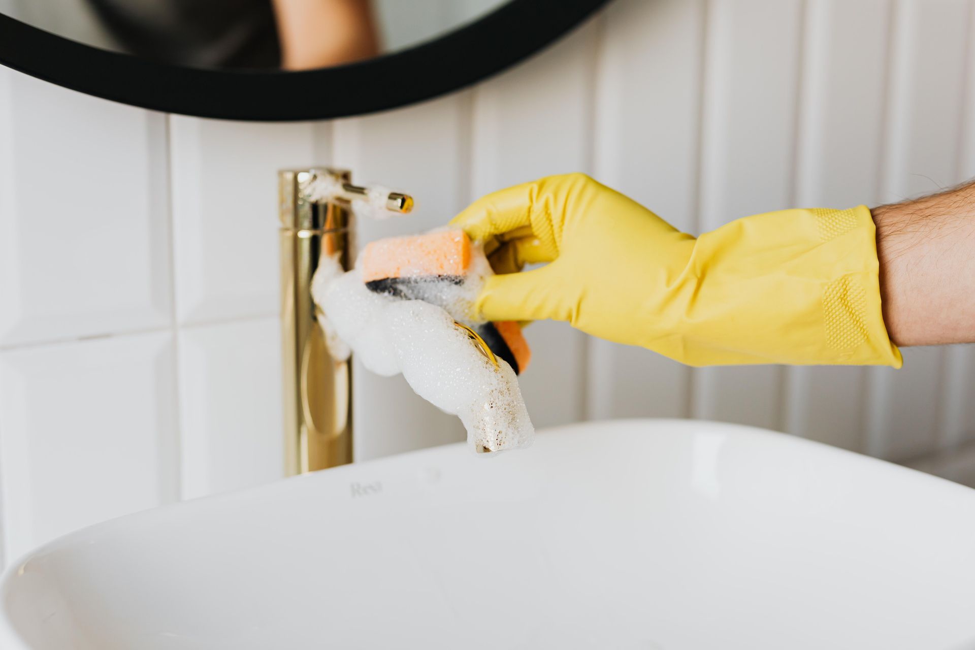 Gloved hand cleaning a gold faucet beside a white sink and mirror