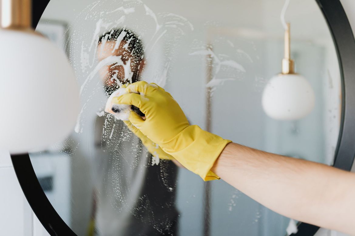 Gloved hand cleaning a mirror with spray cleaner, wiping away smudges on the glass.