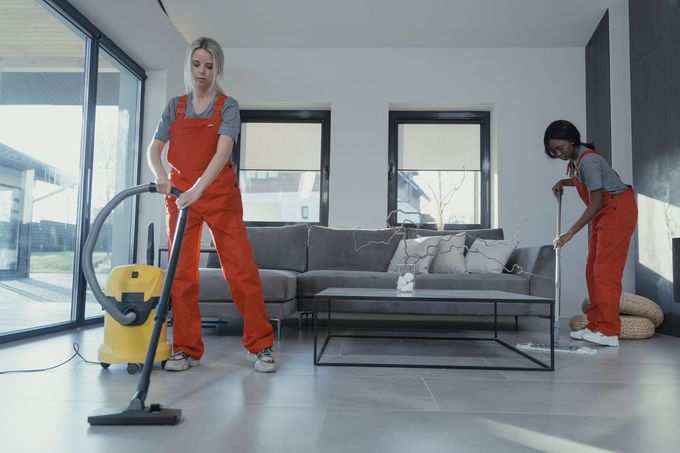 Two cleaners vacuuming a modern living room with a gray sofa and glass coffee table