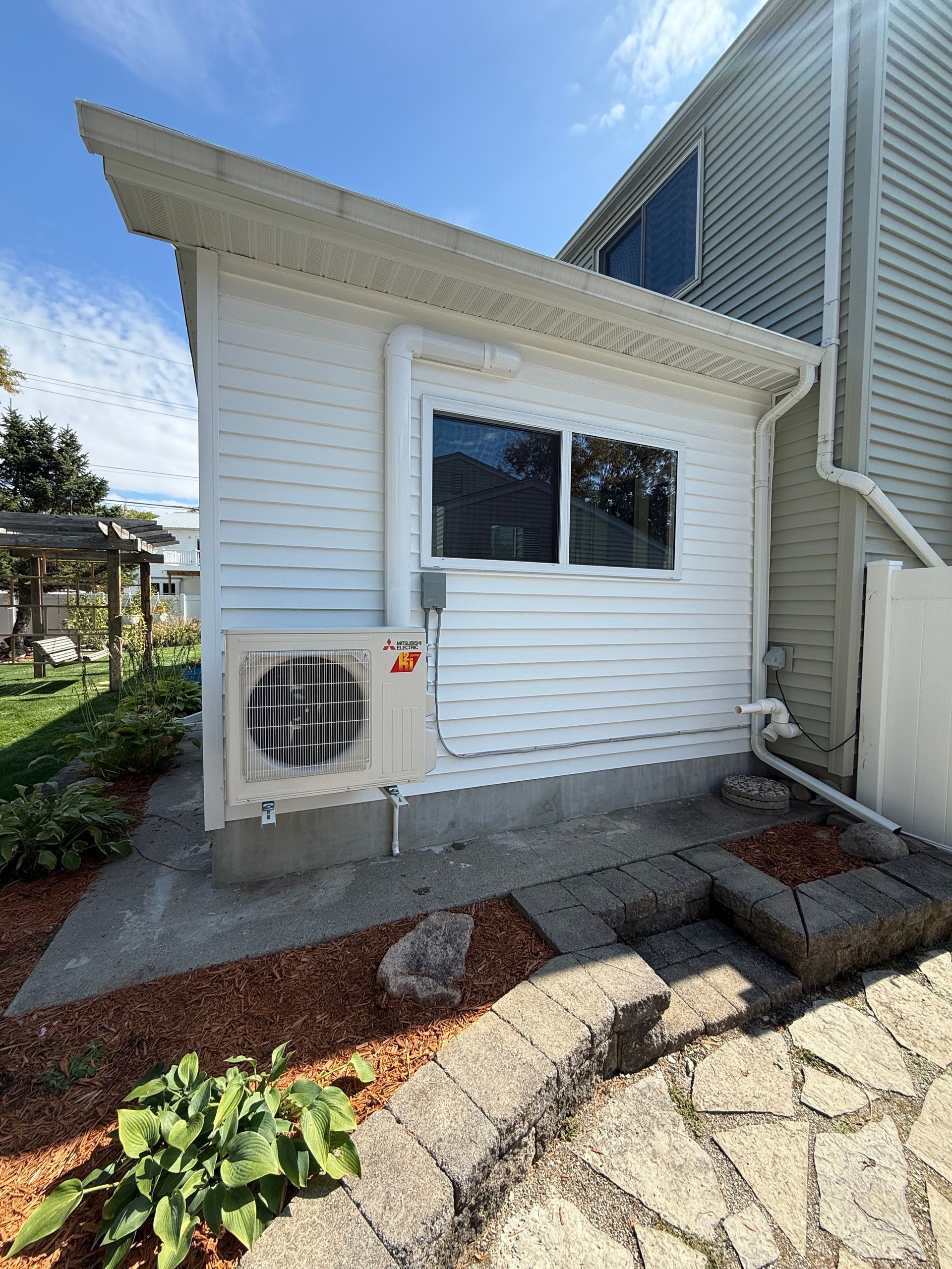 Hand holding gauges, near AC unit. Blue and red gauges, black hoses, white unit outside.