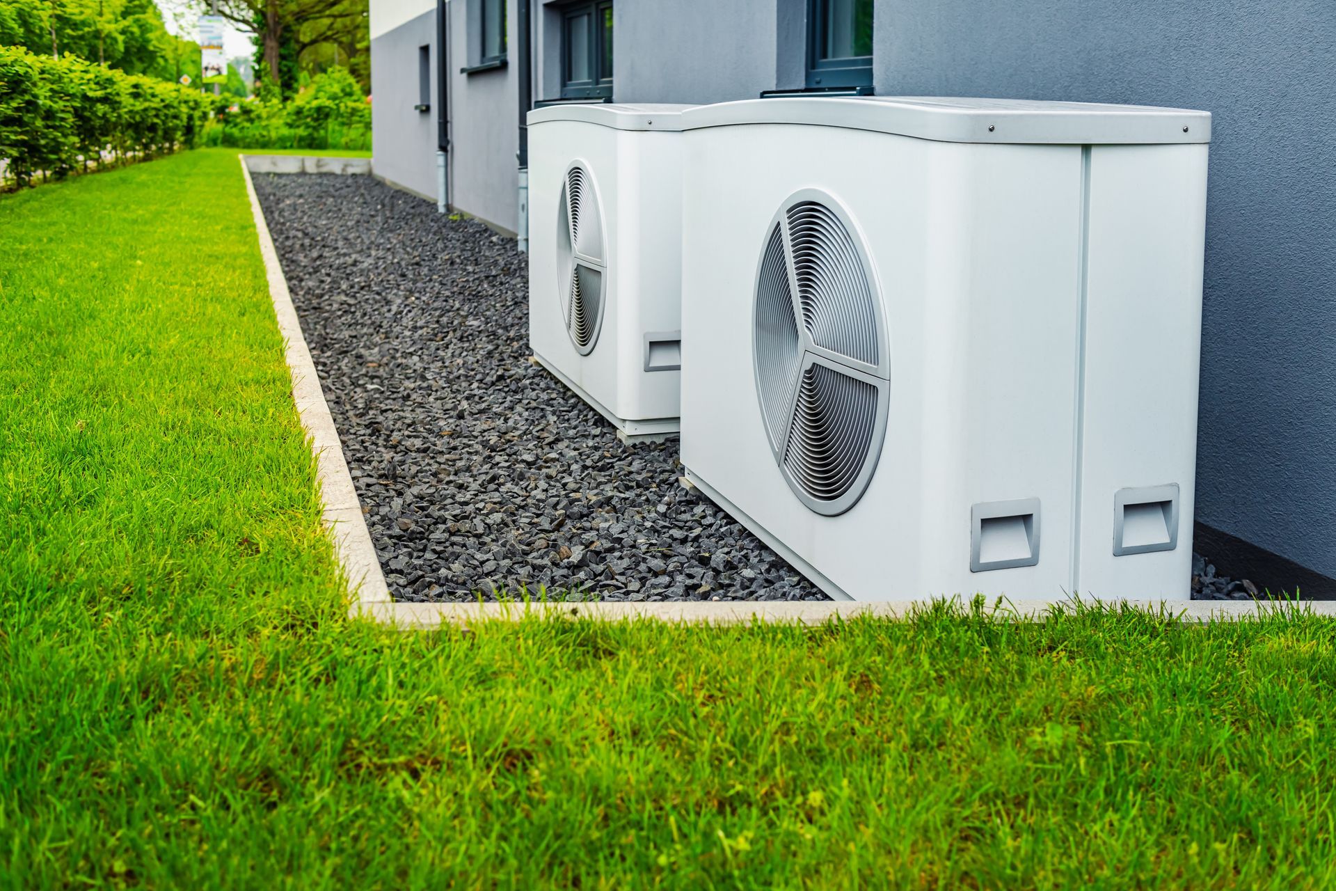 Two white HVAC units on a gravel bed next to a building and lawn.