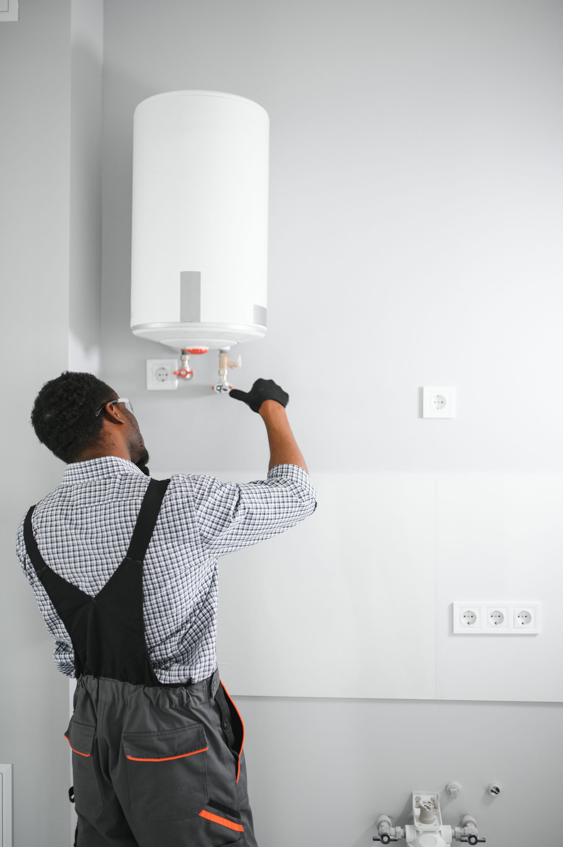 Plumber in gray overalls and gloves works on a water heater, on a white wall.