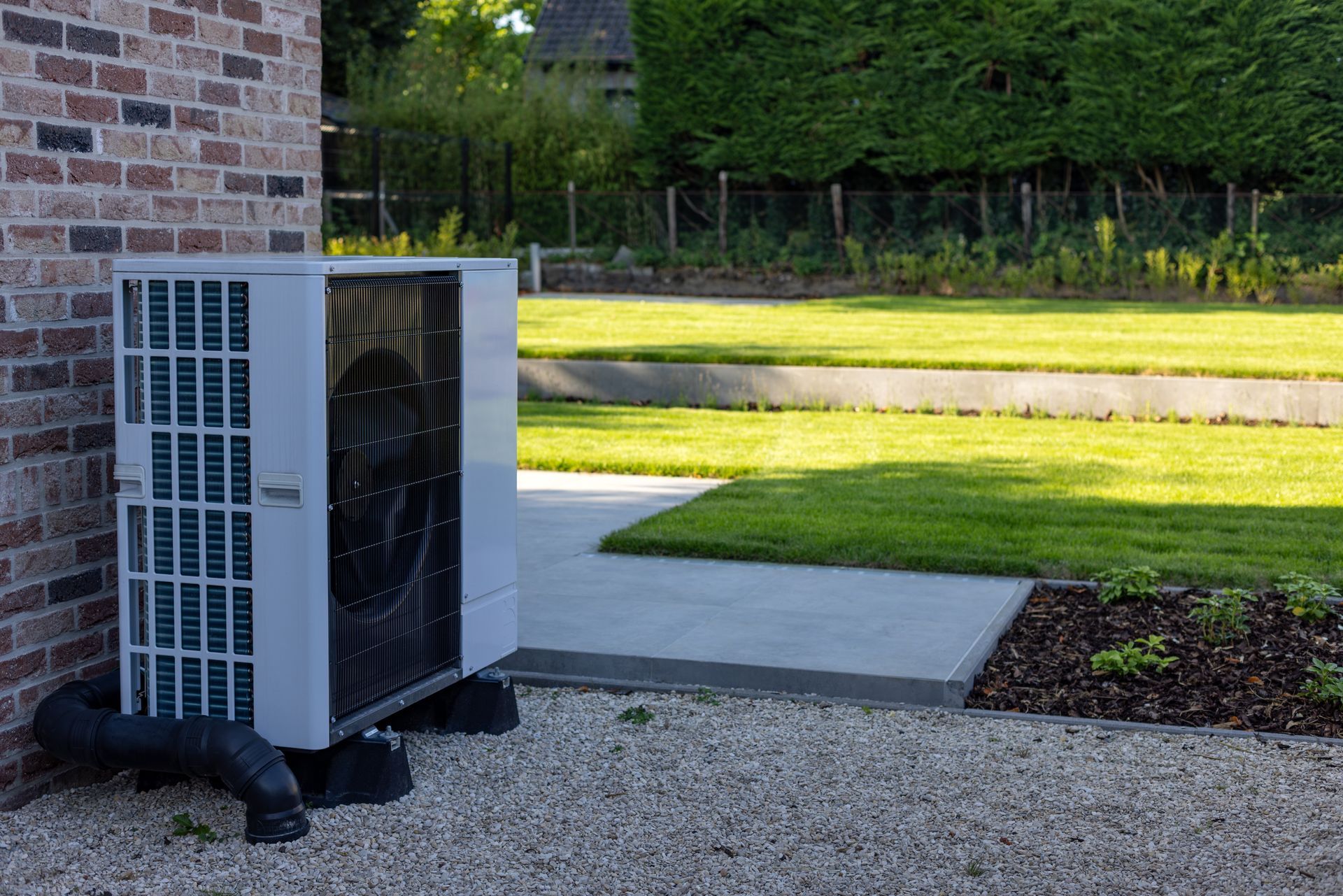 Heat pump unit next to a brick wall and a concrete walkway, with a grassy yard and hedges in the background.