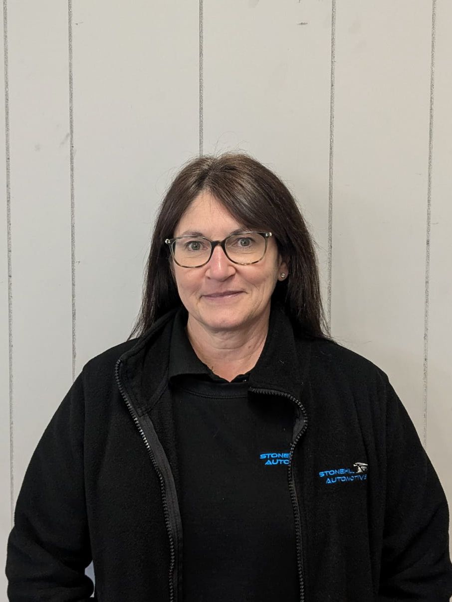 A woman wearing glasses and a black jacket is standing in front of a white wall.