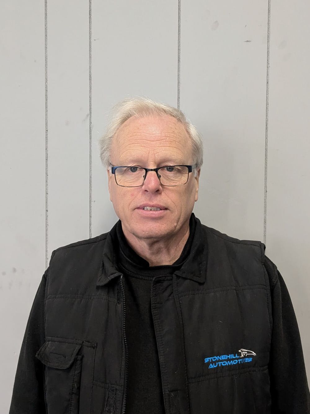 A man wearing glasses and a black vest is standing in front of a wooden wall.