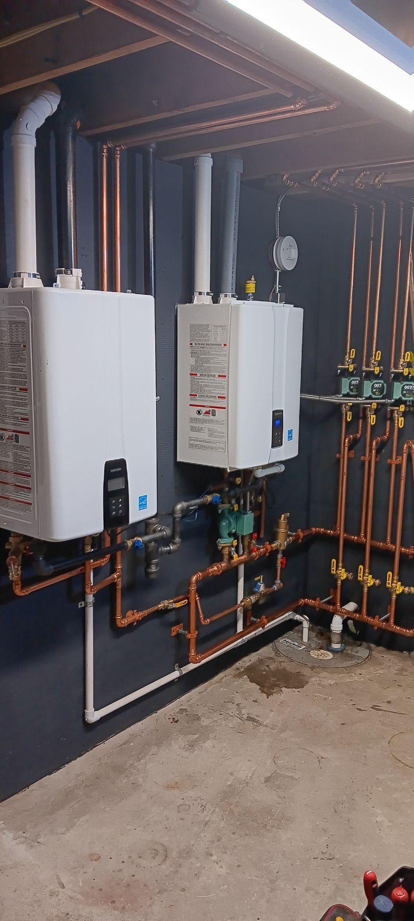 Two white boilers with copper piping against a dark wall in a utility room.