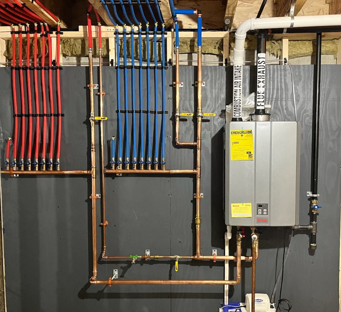 Copper pipes and a tankless water heater installed on a basement wall, with red and blue PEX tubing.