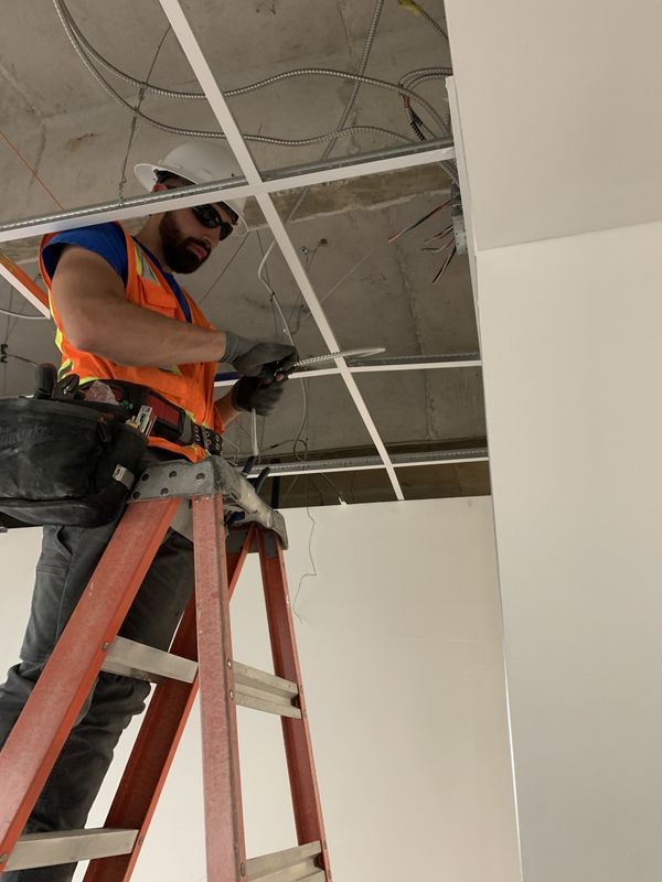 Construction worker on a ladder, installing ceiling components. Wearing safety gear, working indoors with white walls.