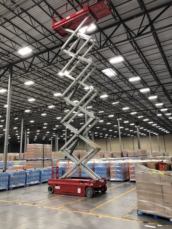 Red scissor lift in warehouse, raised high near the ceiling. Pallets of goods are visible below.
