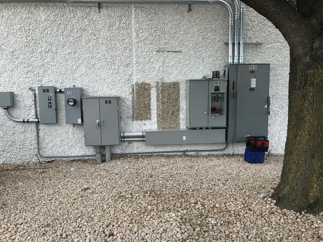 Electrical boxes and conduit mounted on a textured wall near a tree, gravel ground.