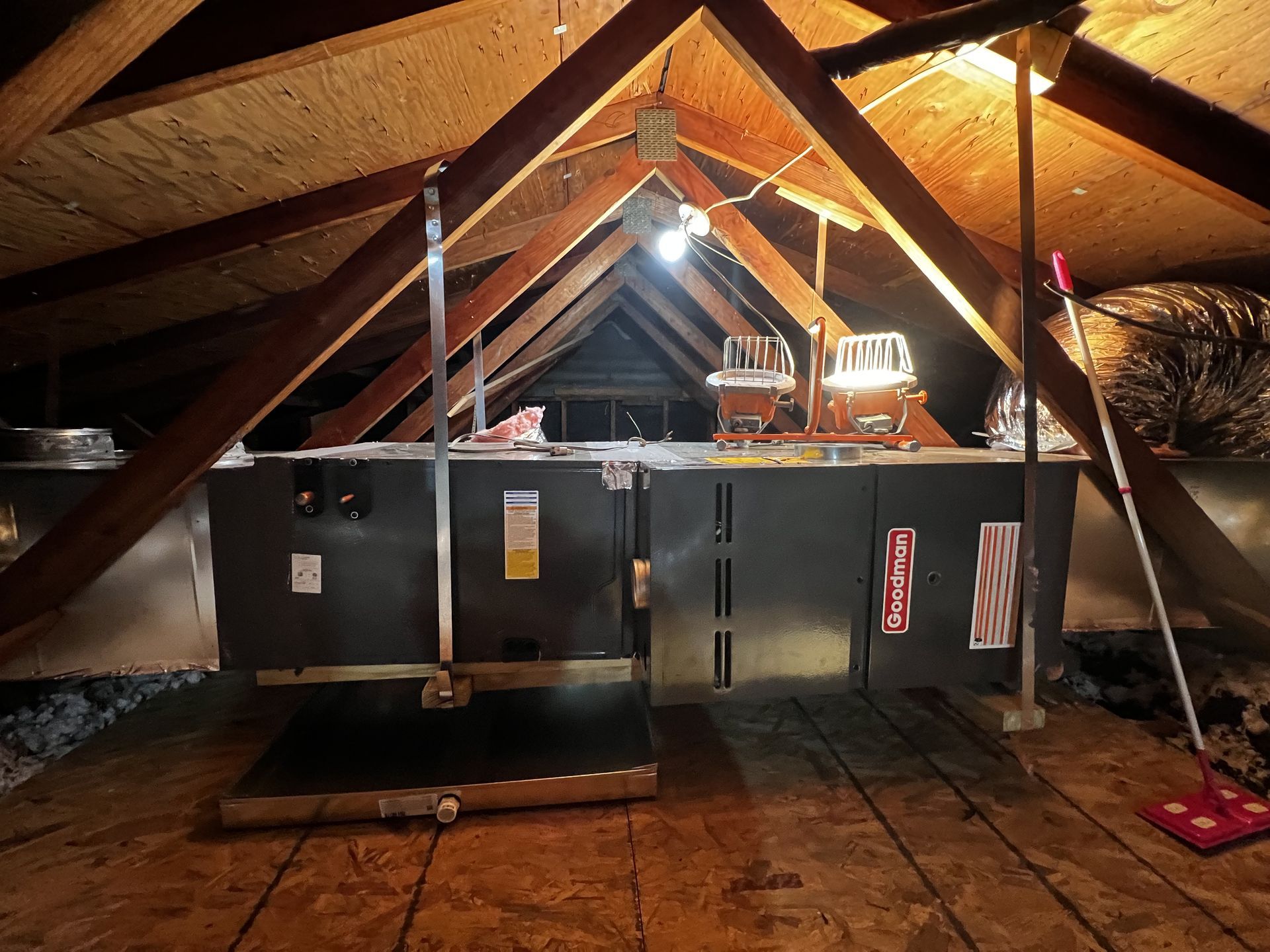 Attic HVAC unit under wooden rafters, with hanging light and insulation on the walls.