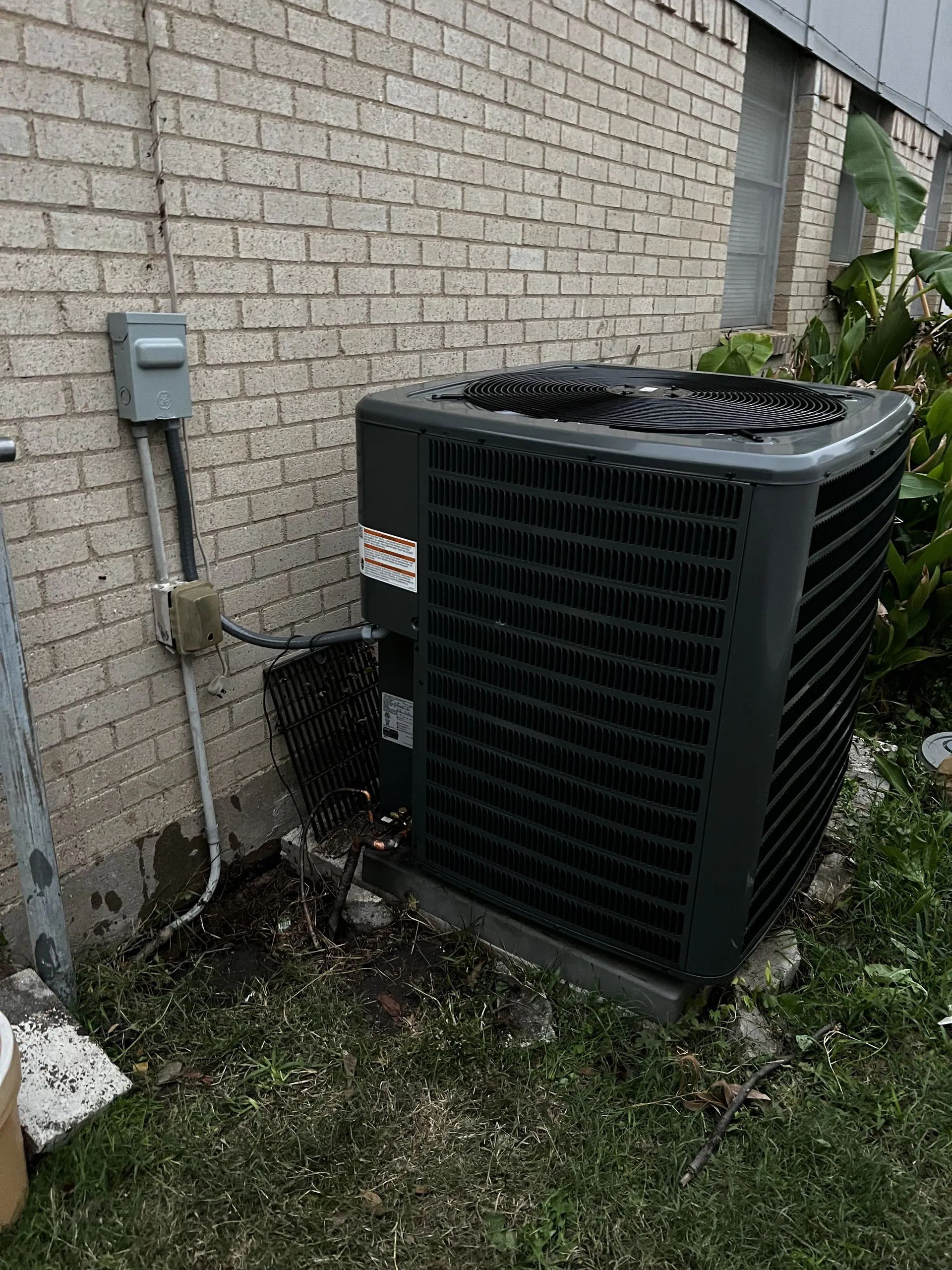 Outdoor HVAC unit beside a brick house wall with electrical meter and grass below