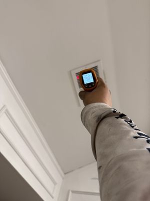 Hand holding a handheld stud finder up to a ceiling near a vent in a white room