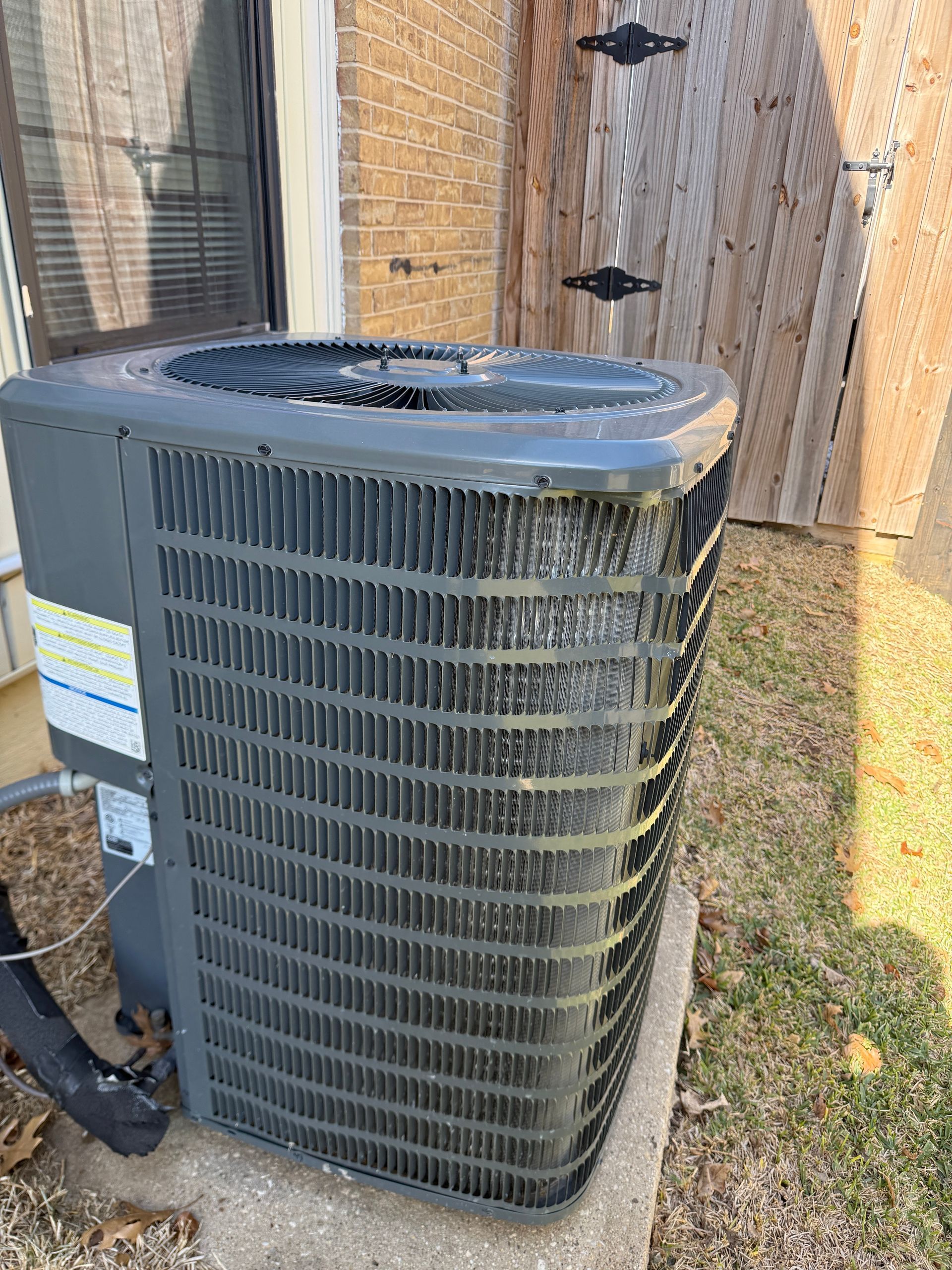 Outdoor HVAC condenser unit beside a wooden fence, with a window and grass nearby.
