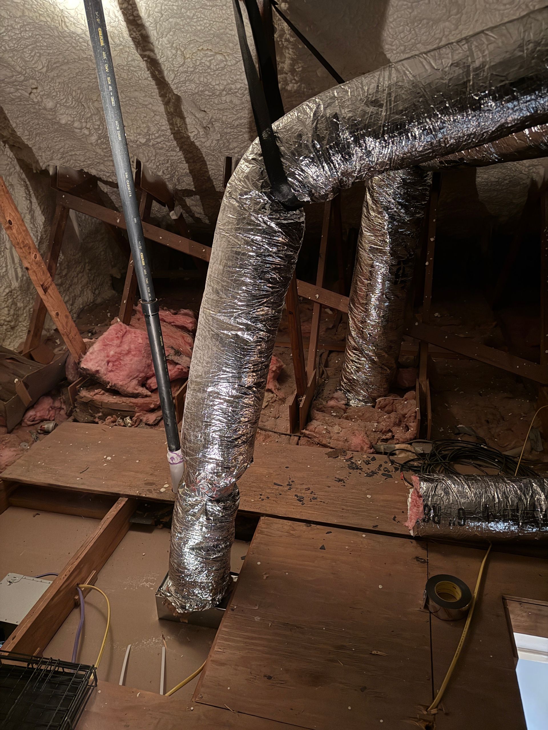 Insulated HVAC ducts and exposed pipes in a dusty attic with damaged flooring and debris