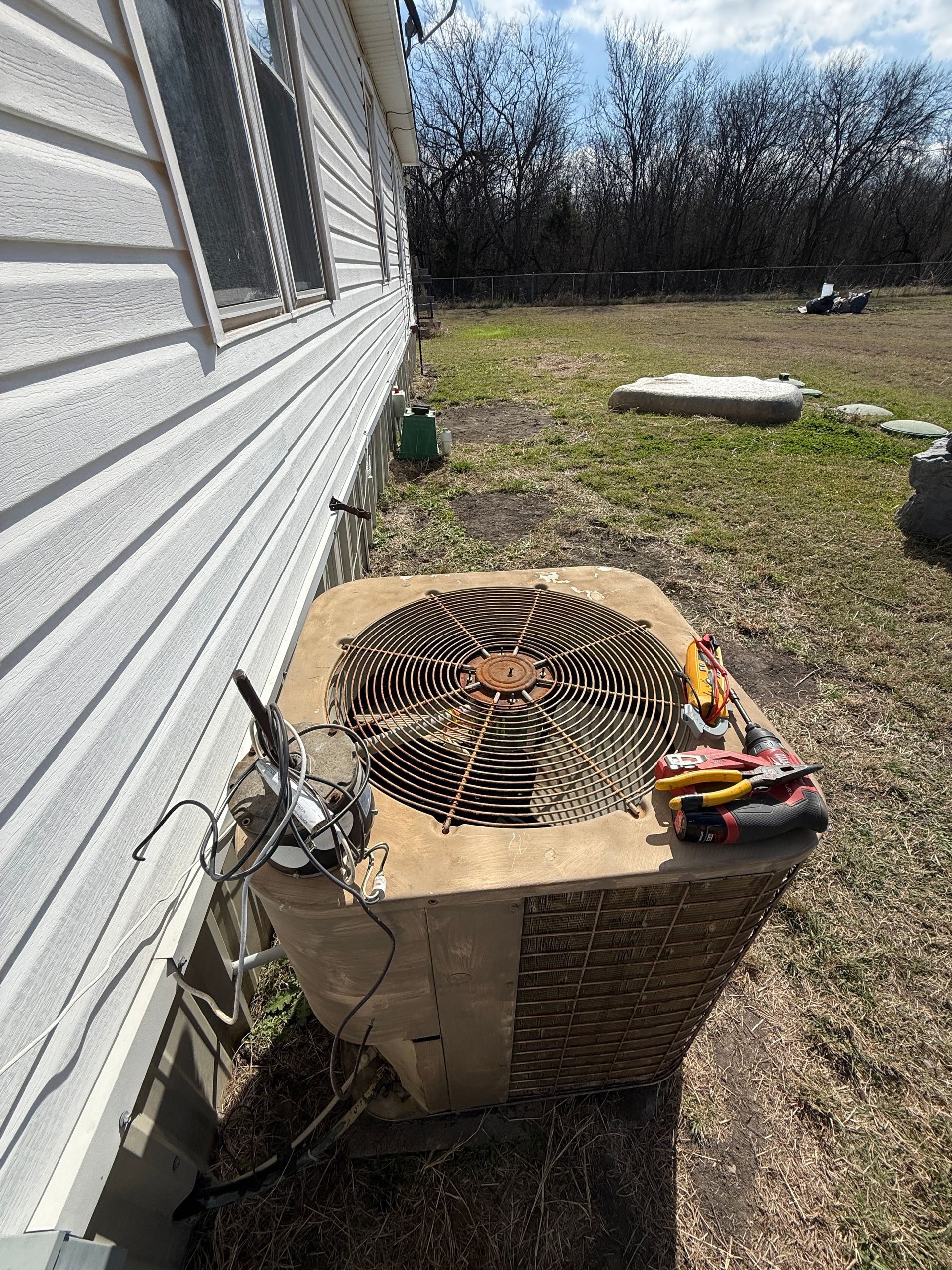 Damaged outdoor air conditioner unit beside a house, with tools on top in a grassy yard.