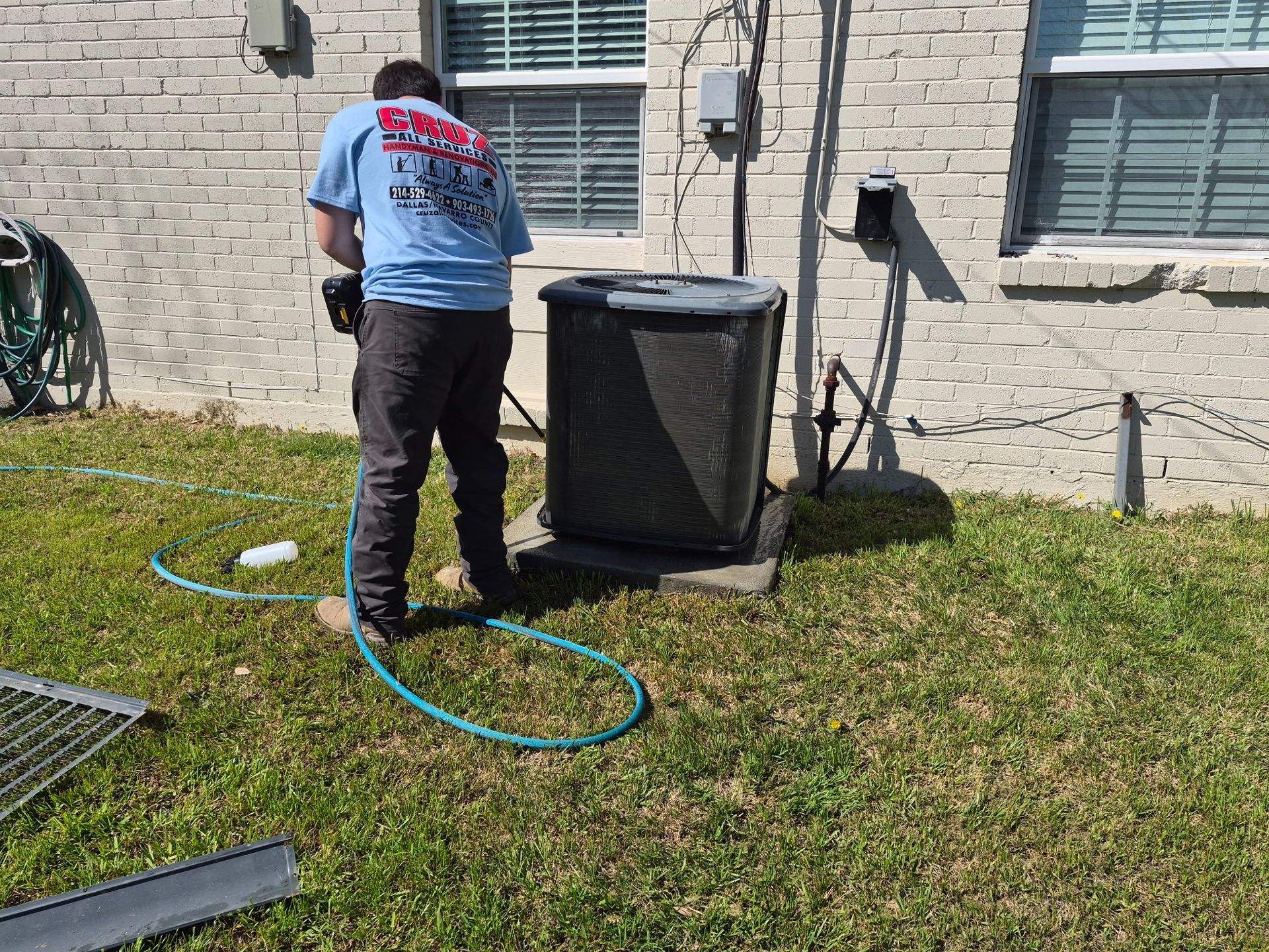 Worker cleaning an outdoor HVAC unit beside a brick house with a hose.