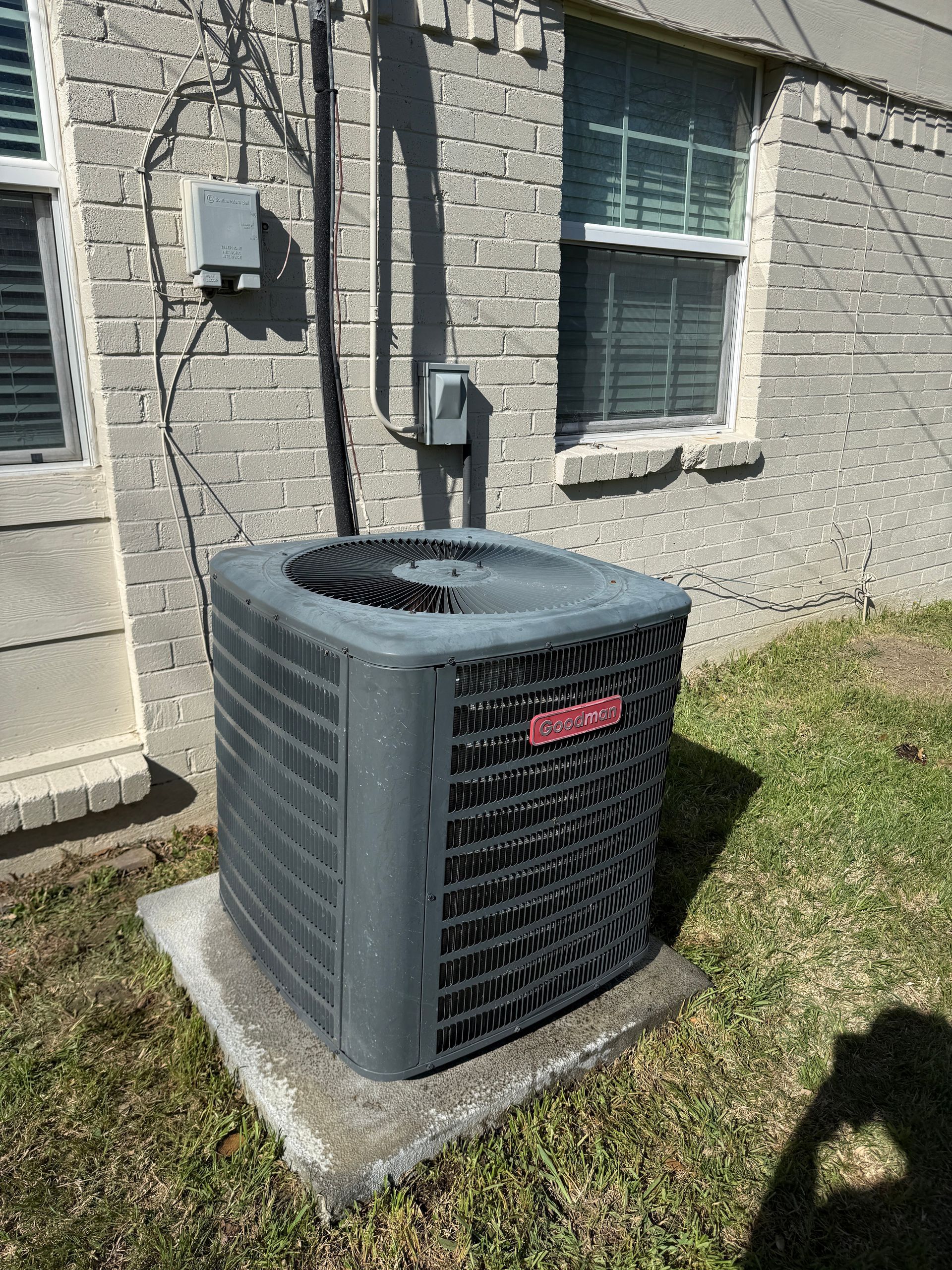 Outdoor air conditioning unit beside a beige house wall and window on a concrete pad.