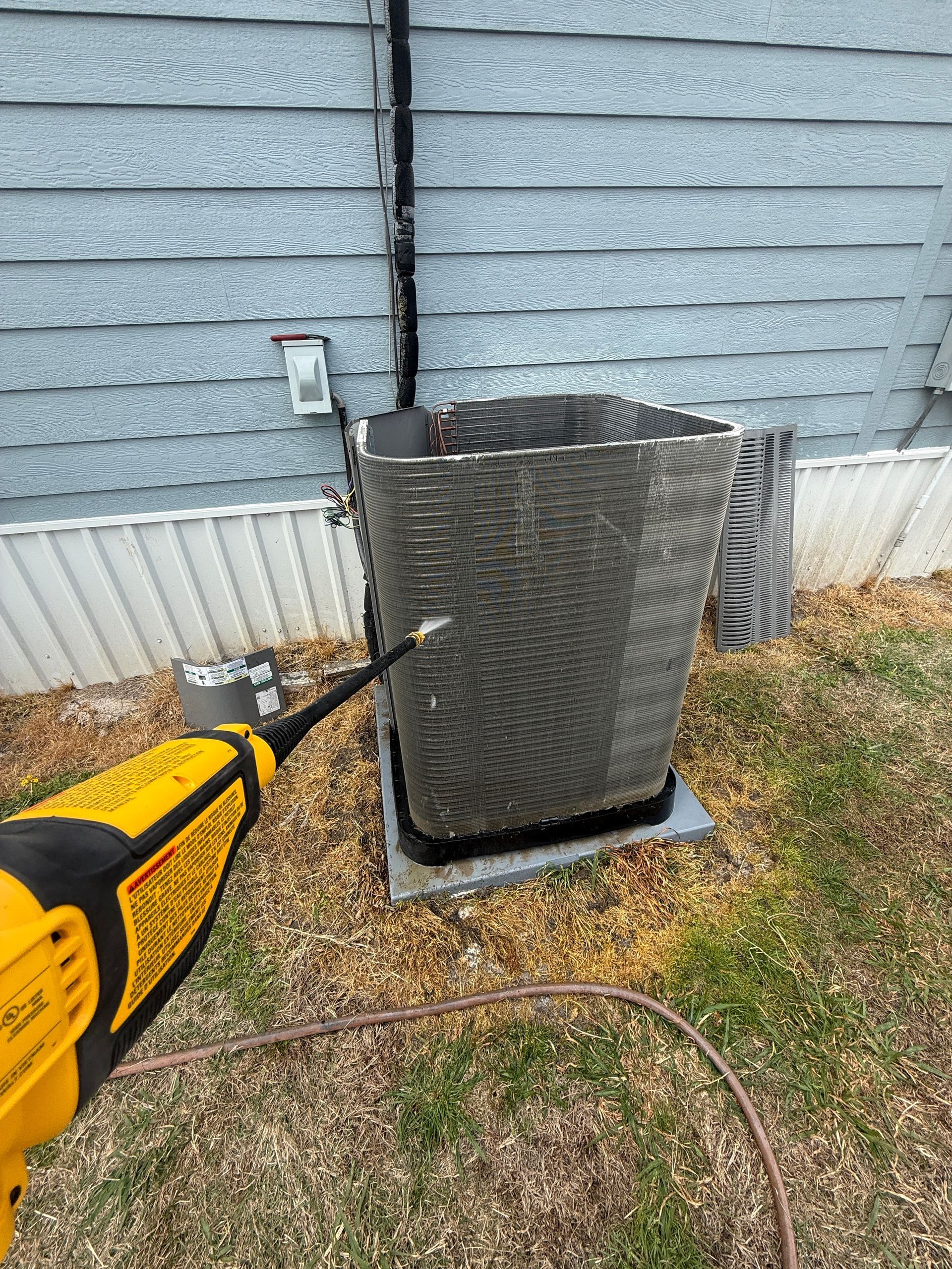 Yellow pressure washer spraying an outdoor HVAC unit beside a house siding.