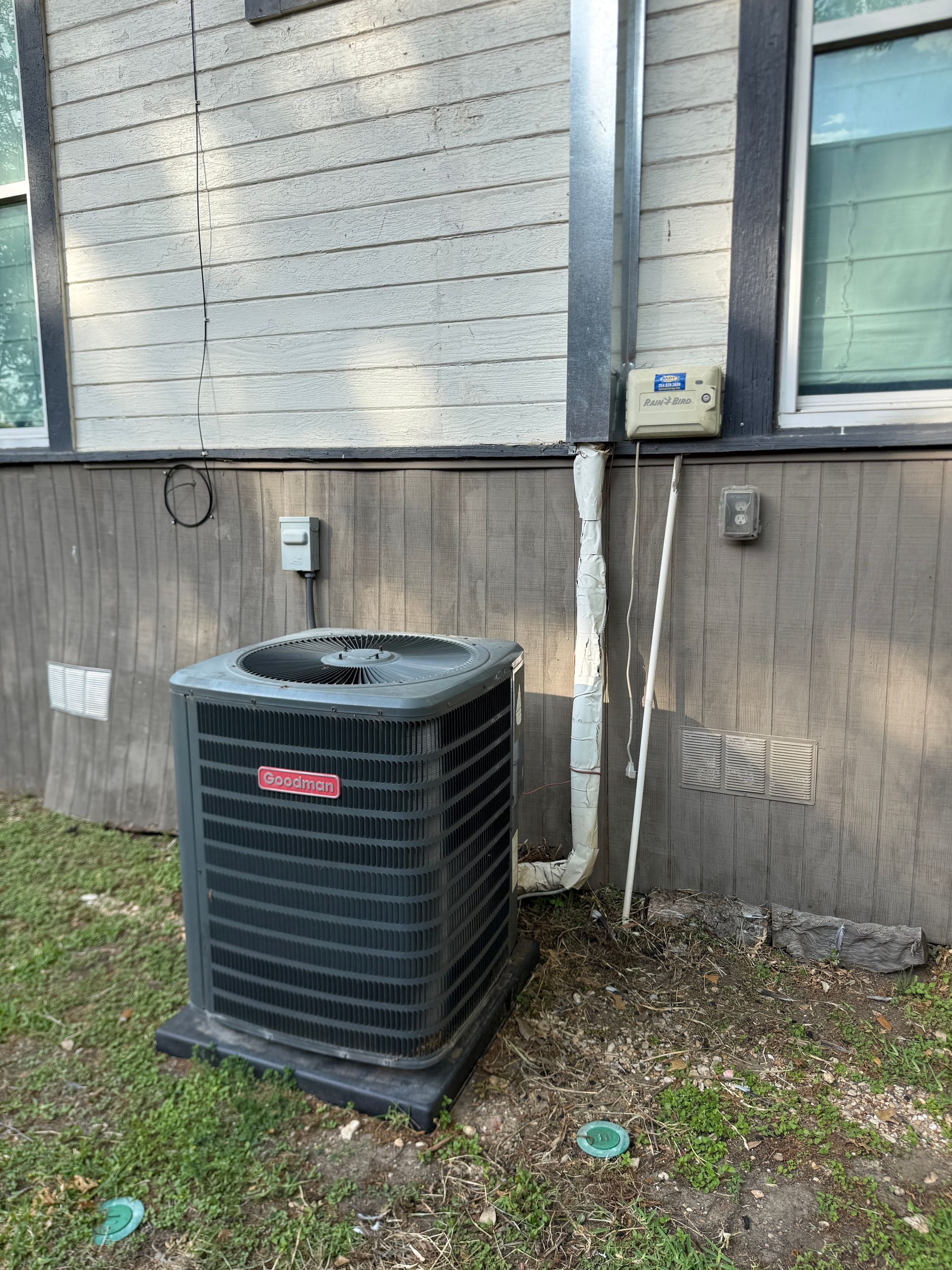 Outdoor air conditioner unit beside a house wall and utility meters, with grass and dirt around it