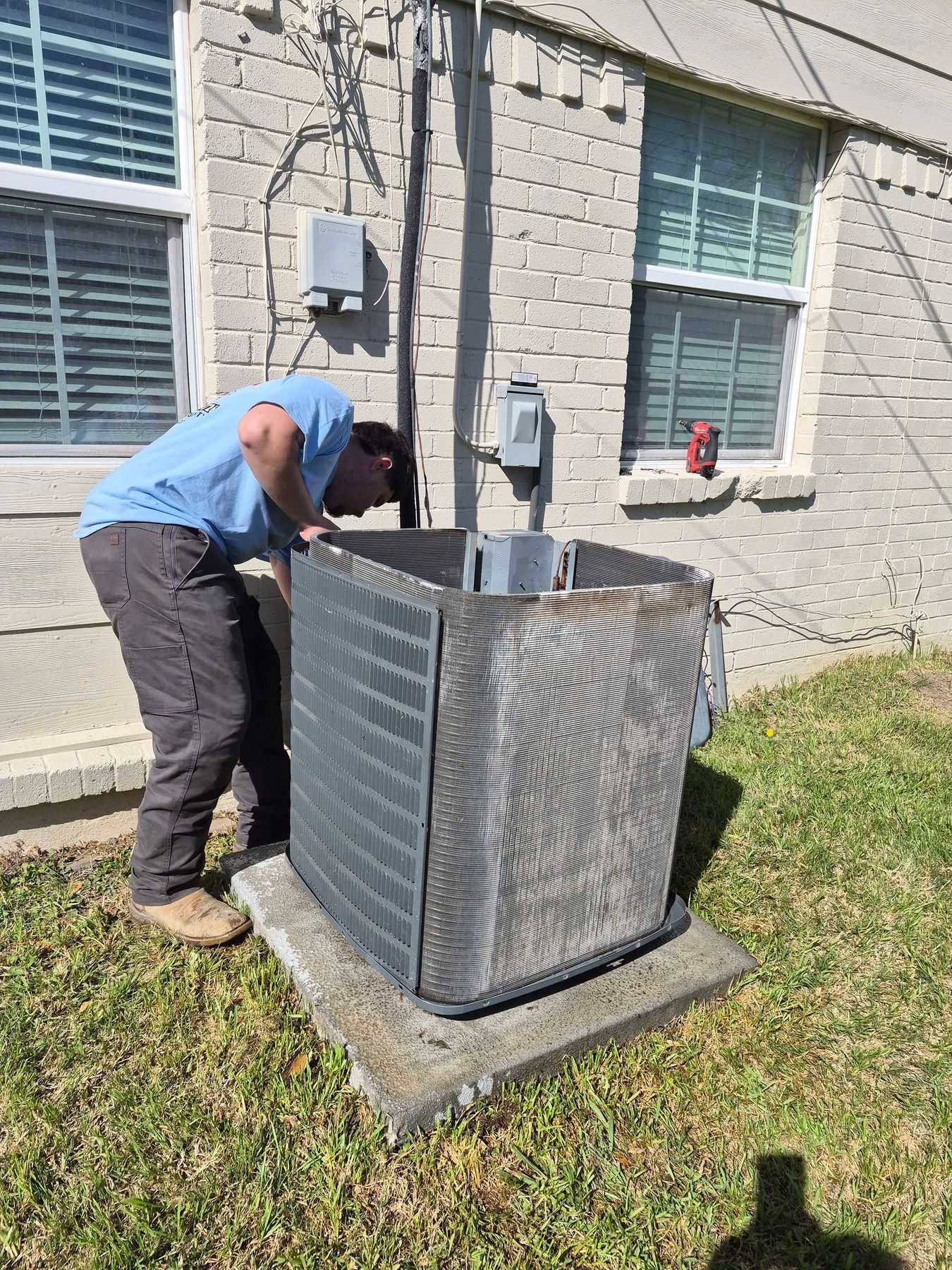 Worker servicing an outdoor HVAC unit beside a house window, leaning over the open top panel.
