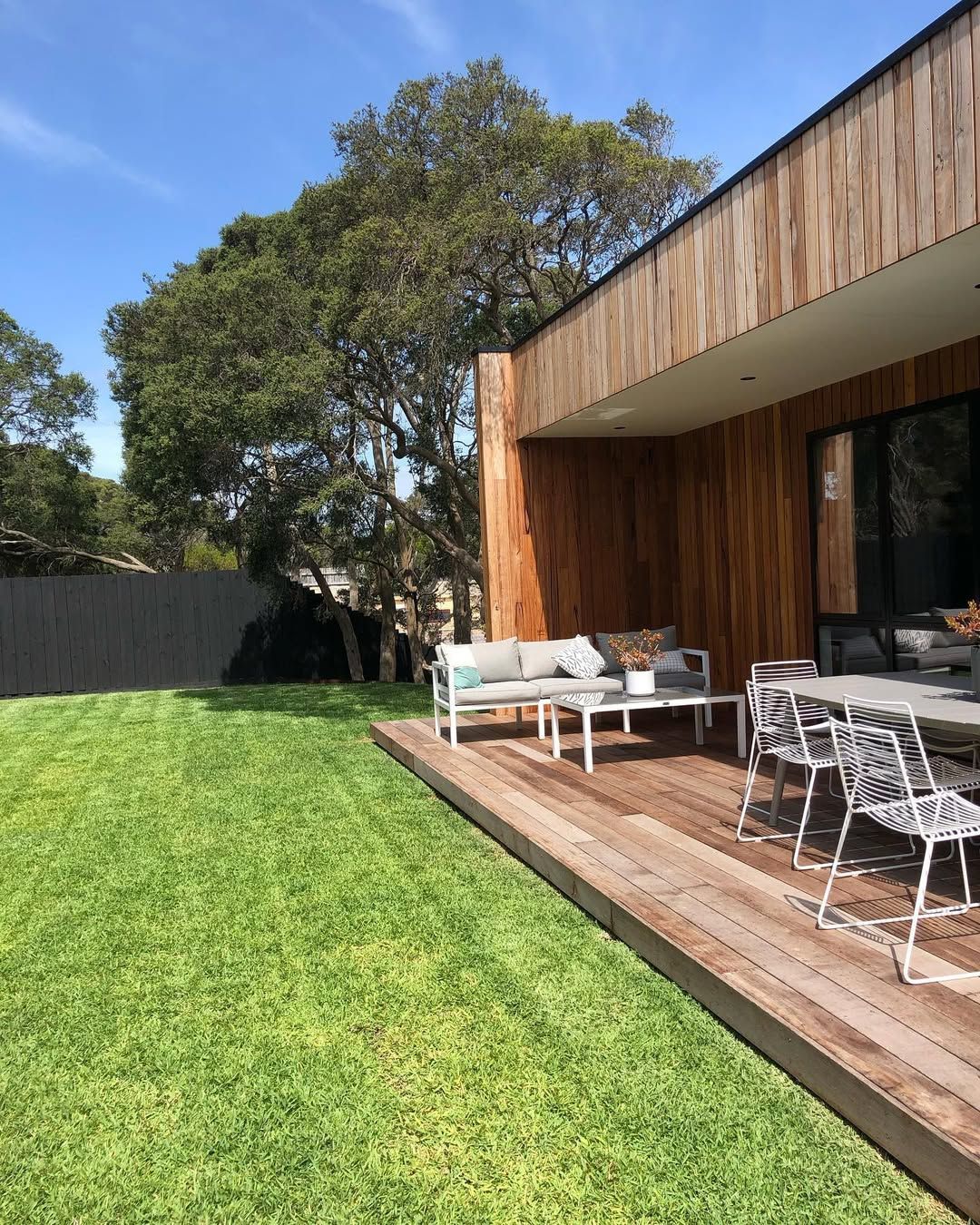 Green lawn, wooden deck and building exterior. Outdoor seating on deck, lush tree in background, bright blue sky.