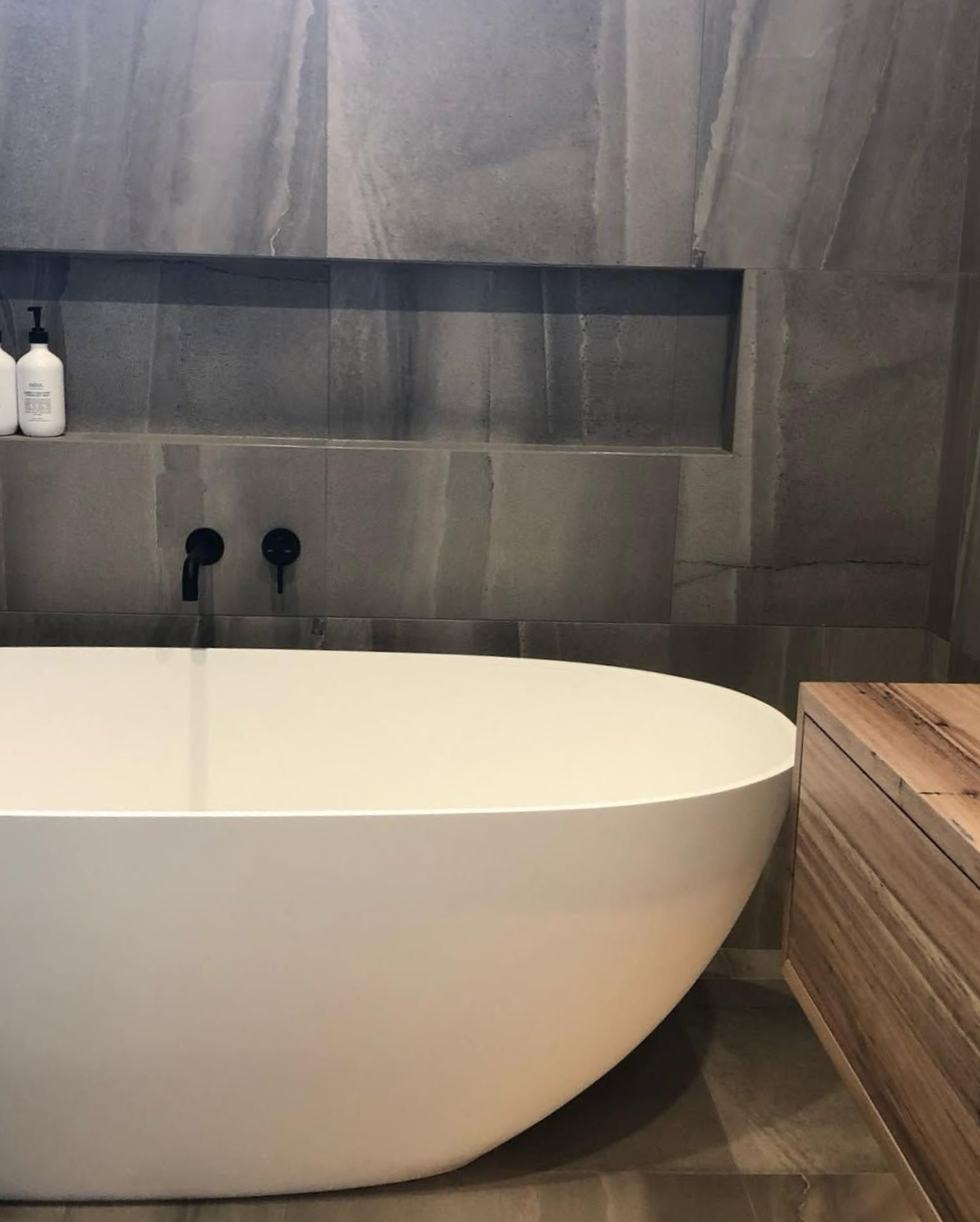 Modern bathroom with white oval tub, gray tile walls, and wooden cabinet.