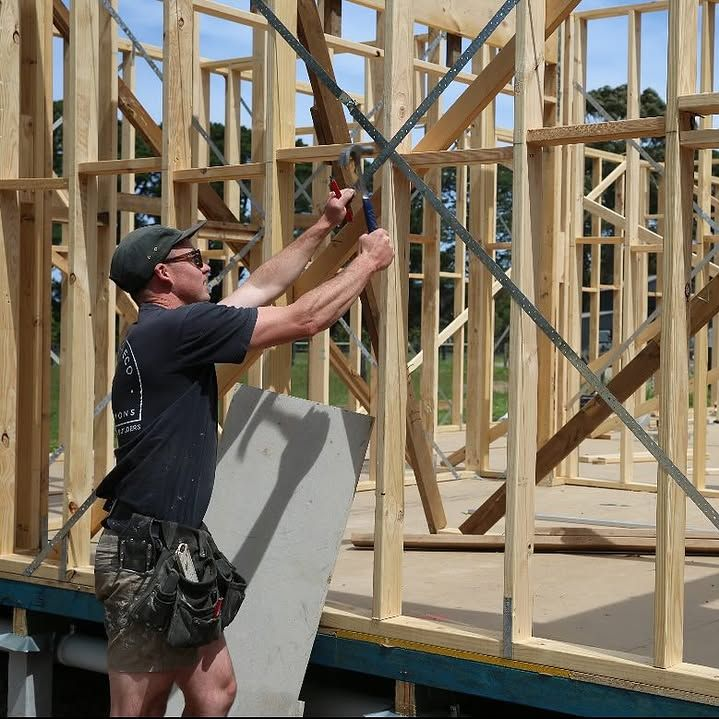 Man using pliers on metal bracing in a wooden house frame under construction.