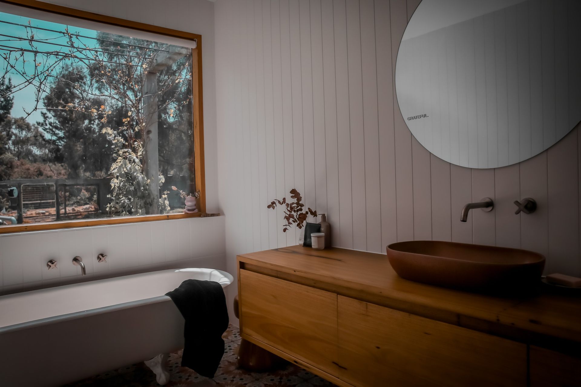 Bathroom with a white clawfoot tub, wooden vanity, round mirror, and large window with trees.