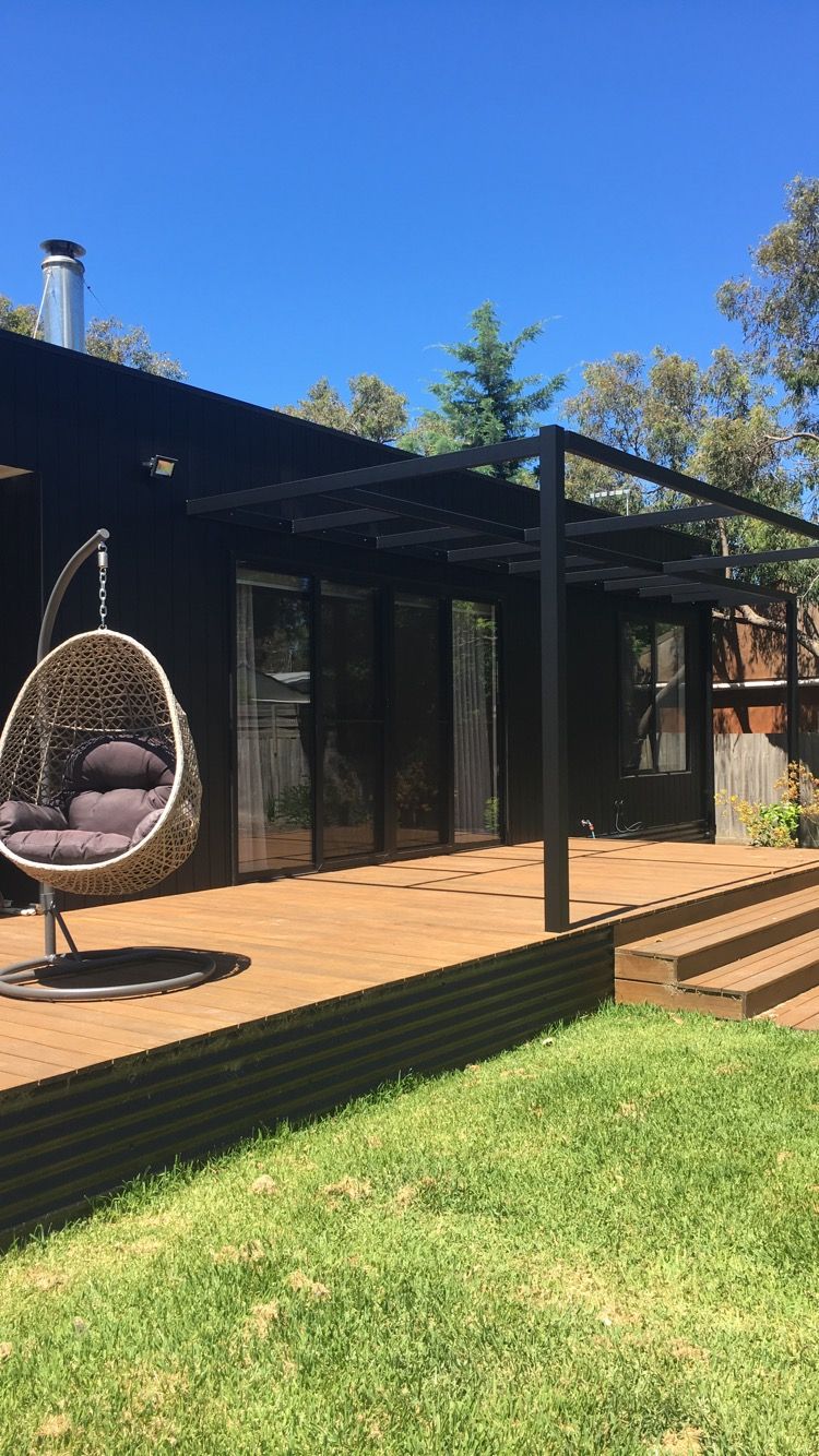 Black house with wooden deck, pergola, and hanging chair. Green grass and blue sky.