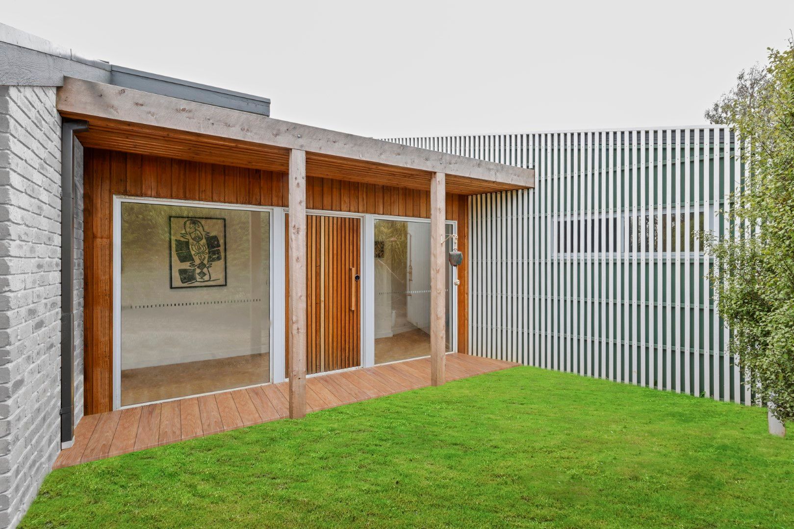 A wooden-framed outdoor room with glass doors opens onto a green lawn, and next to a slatted white wall.