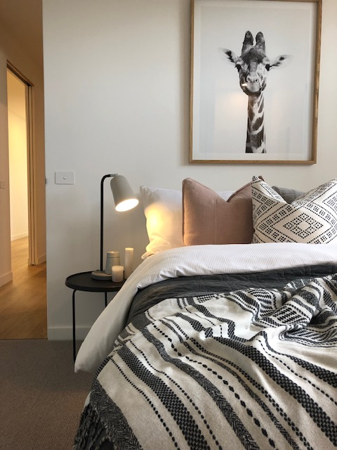 Bedroom with giraffe art, bedside table with lamp and candles, patterned blanket on bed.