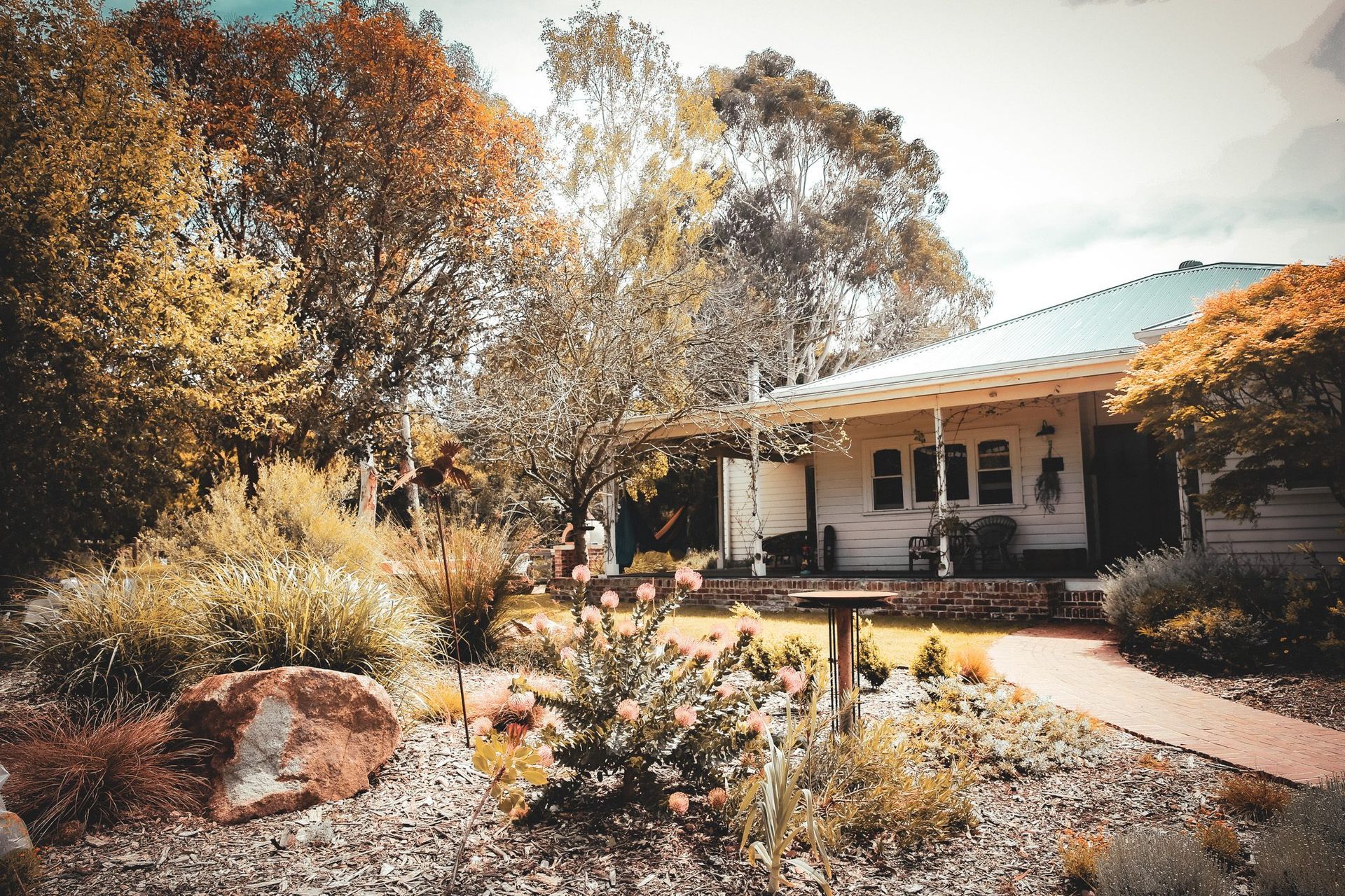 White house with porch surrounded by garden with trees and shrubs.