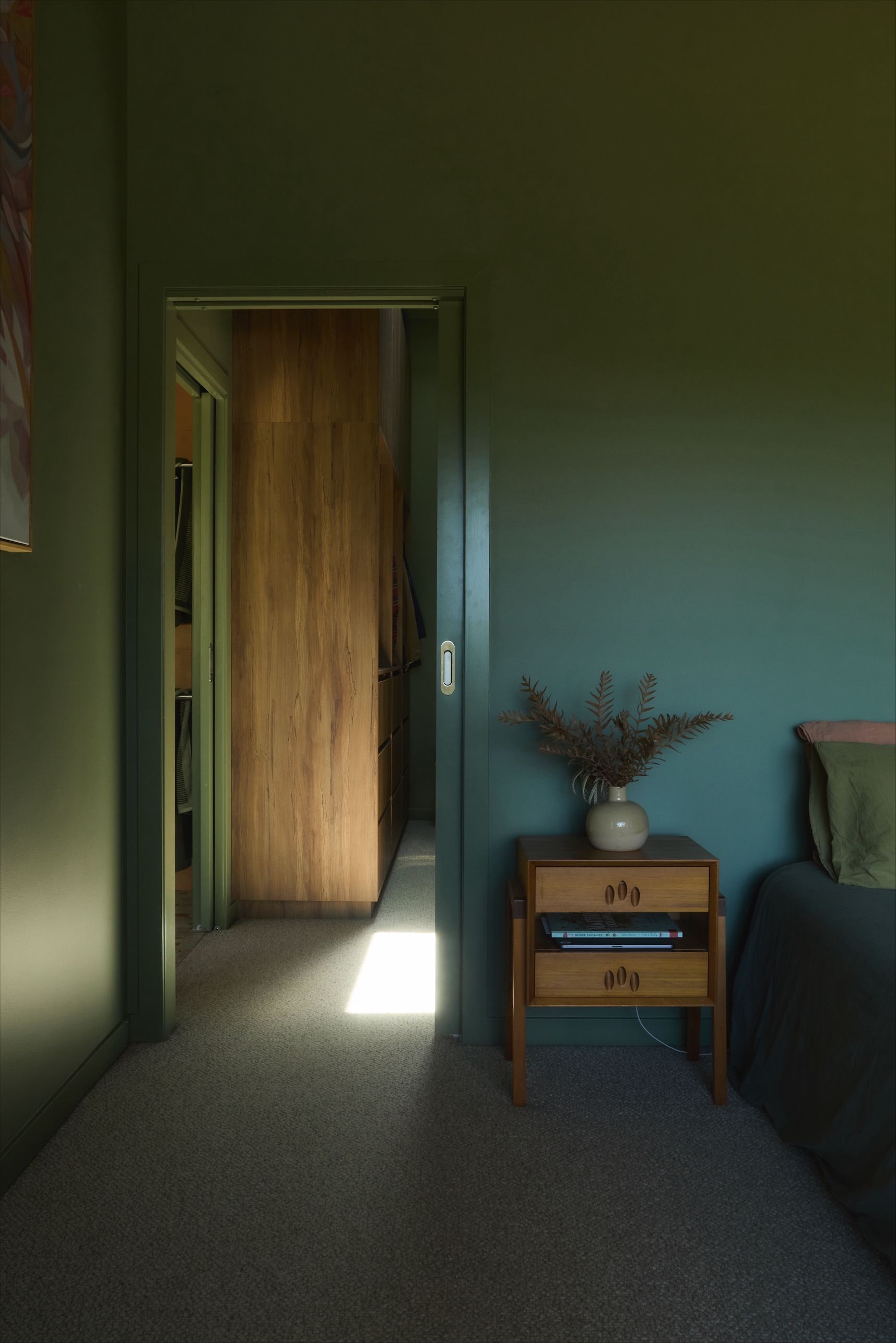 Green-walled bedroom with open doorway to a hallway with a wooden closet and sunlight streaming through. A nightstand sits next to a bed.