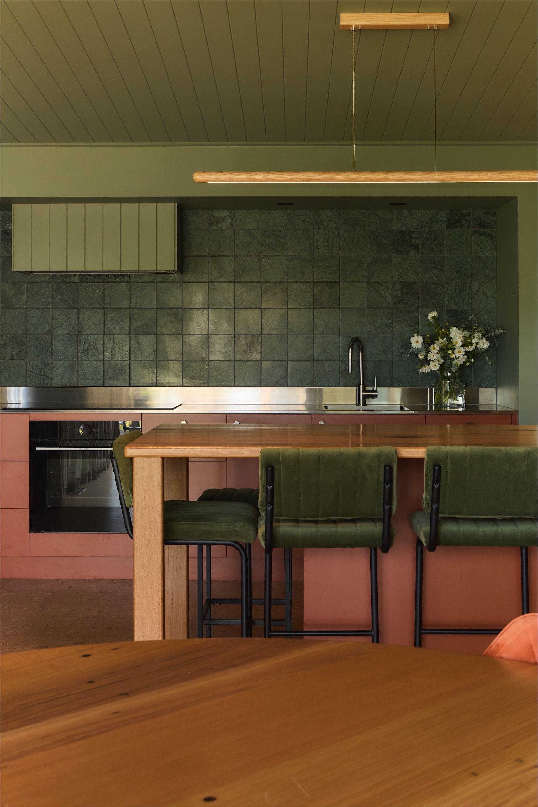 Modern kitchen with green tile backsplash, pink cabinets, and wooden island with green chairs.