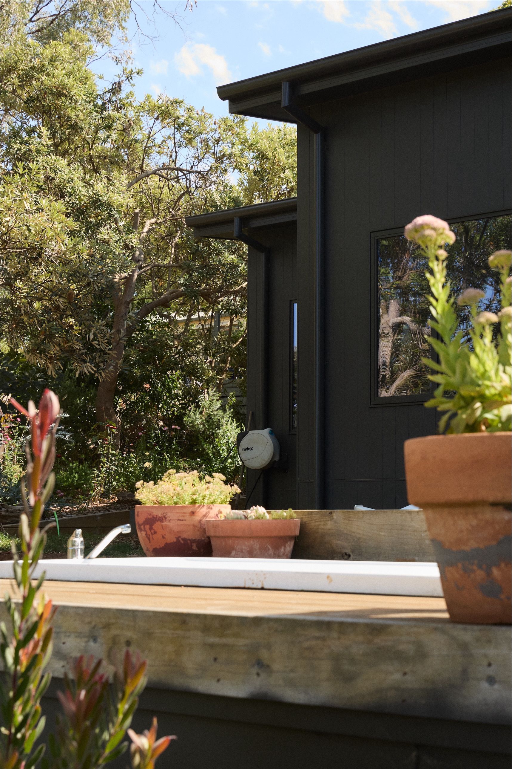 Black building exterior with potted plants on a concrete ledge, trees in background, sunny day.