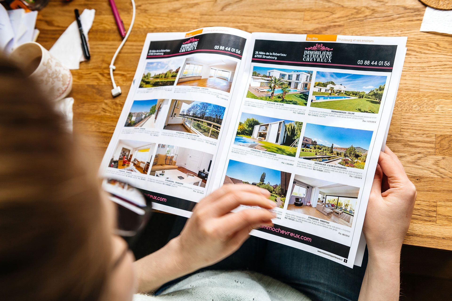 Hands holding an open brochure displaying various property listings on a wooden desk.