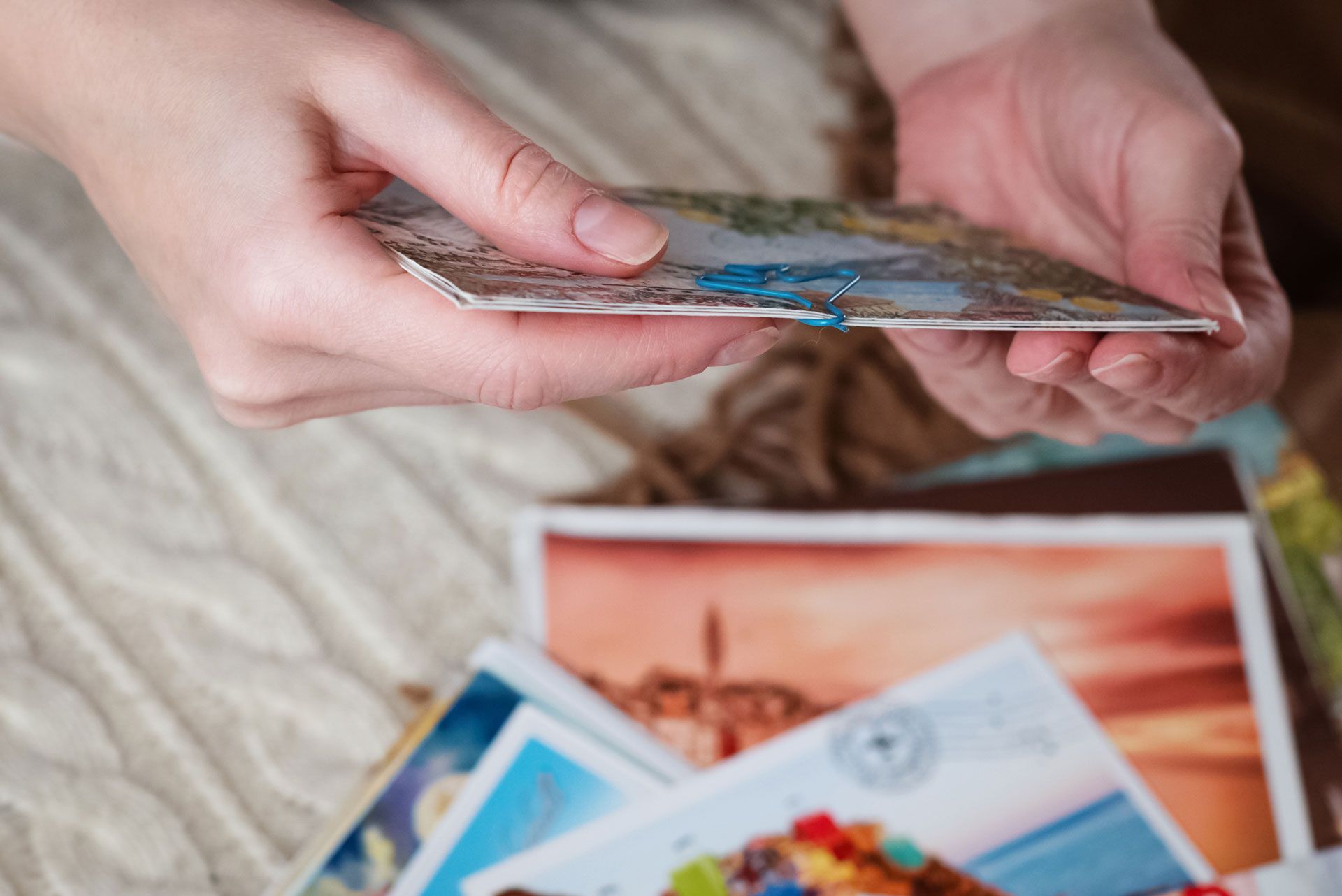 Hands hold a stack of colorful postcards above a collection of other photos on a textured surface.