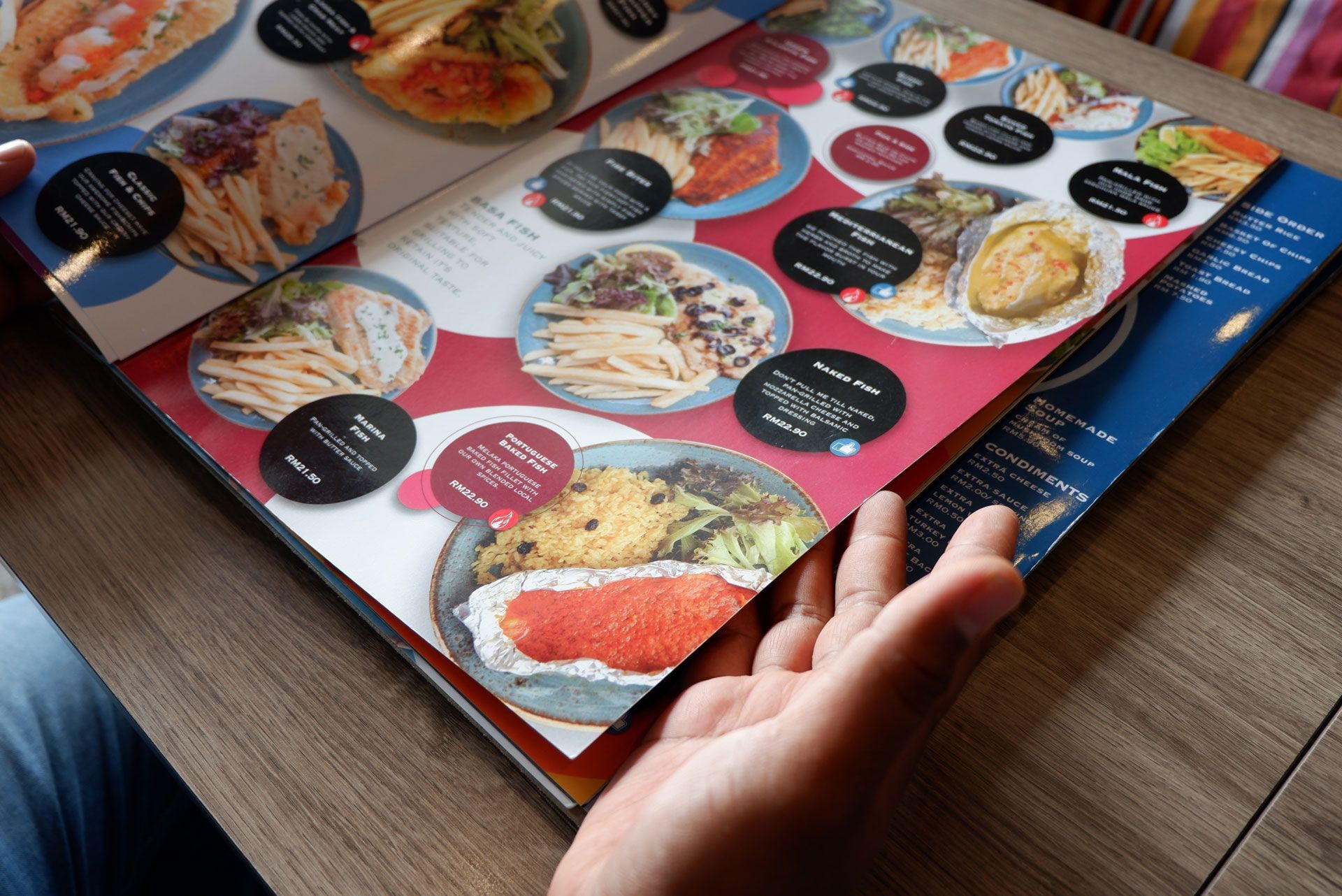 A person’s hand holds open a restaurant menu featuring photos of various meals against a red and blue graphic background.