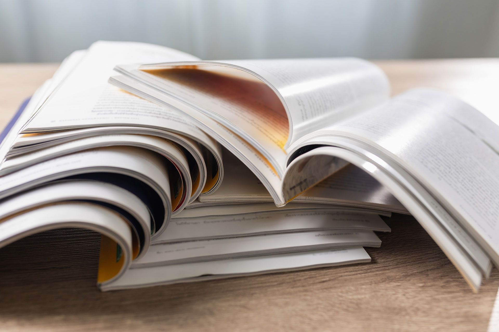 A close-up of several open, layered magazines stacked on a wooden surface, with pages fanned out.