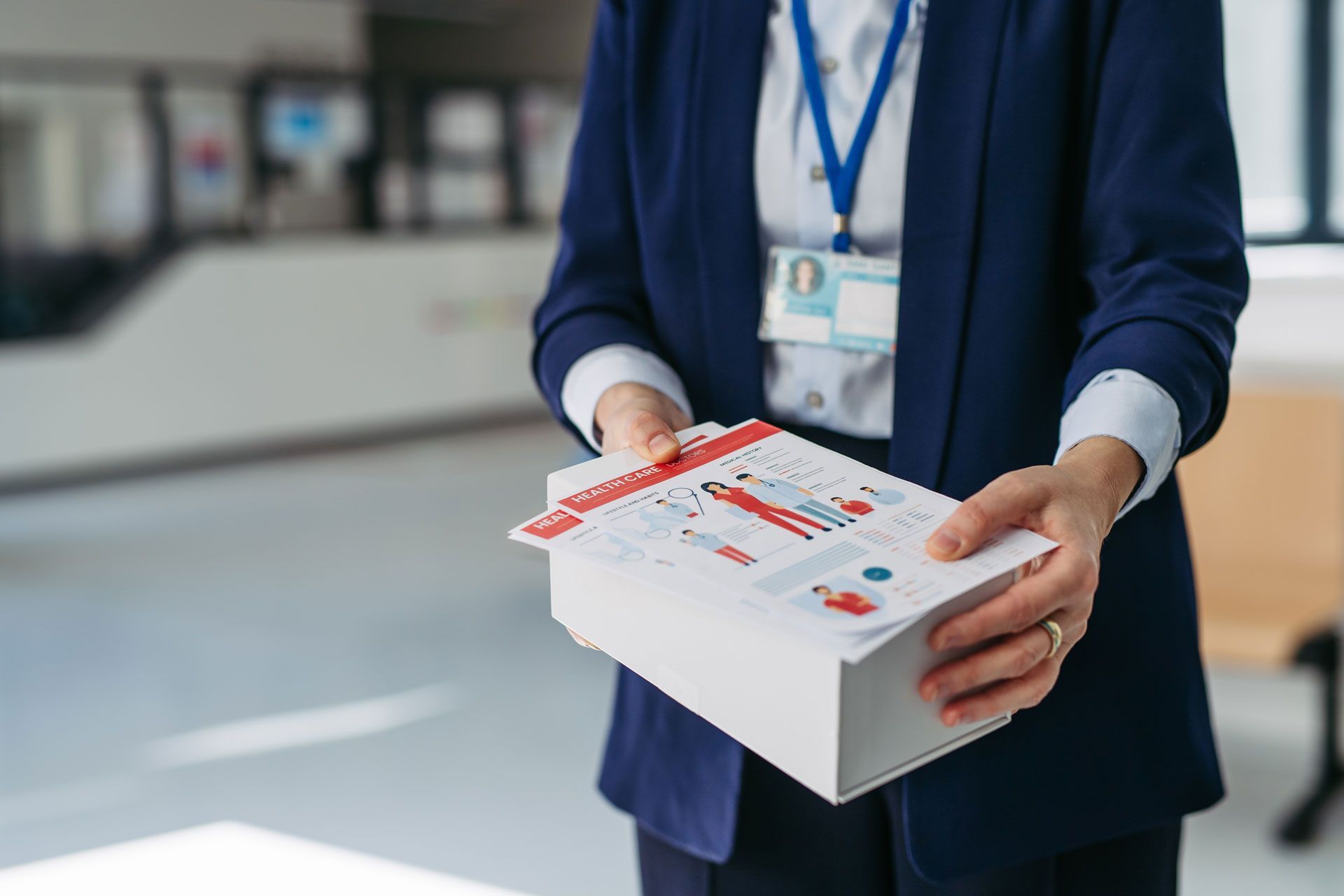 A person in a blue blazer holds a stack of white informational booklets featuring red and blue graphics in a bright office.