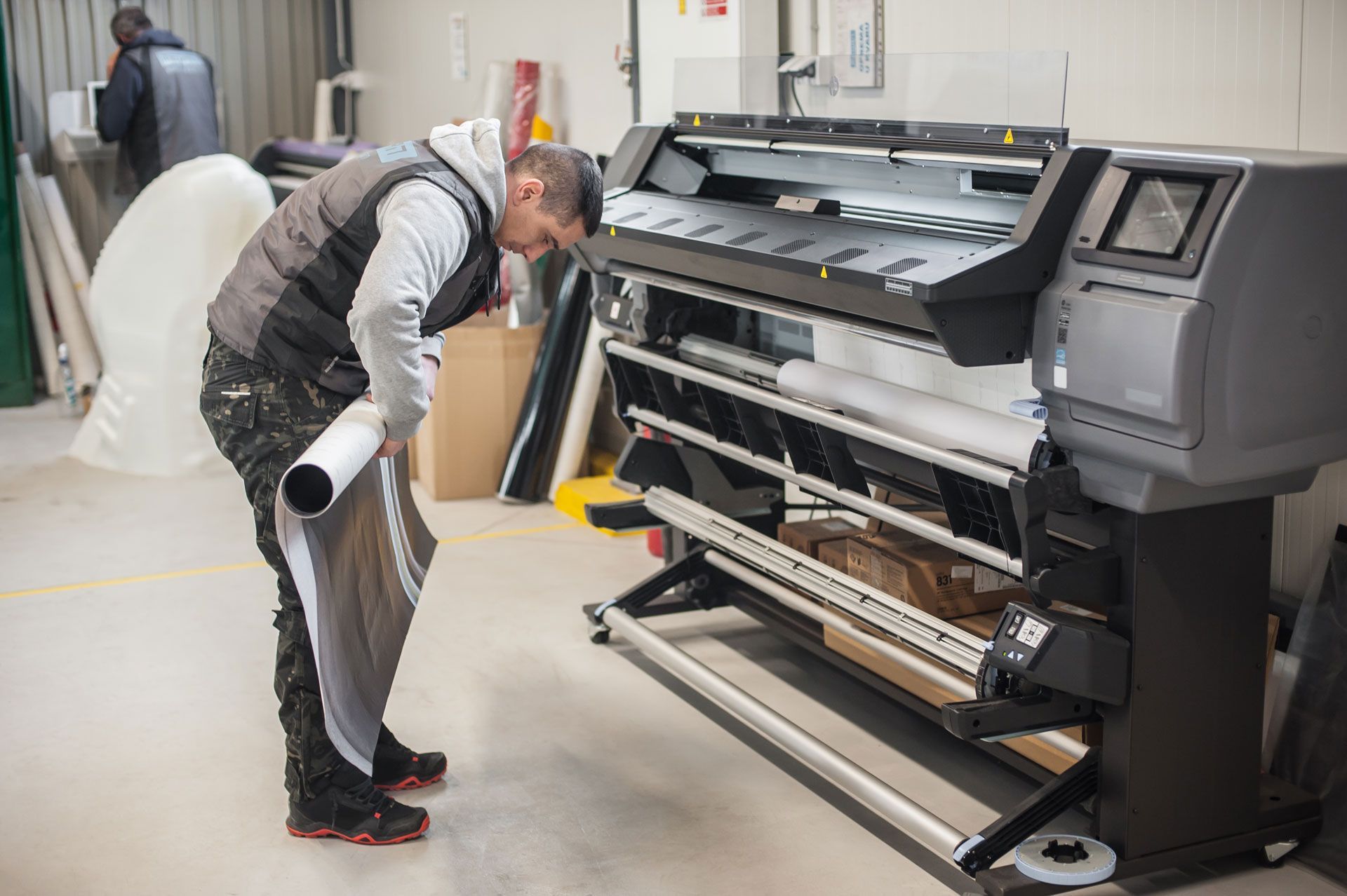 A person in a gray vest and camo pants loads a roll of printing material into a large, industrial wide-format printer.