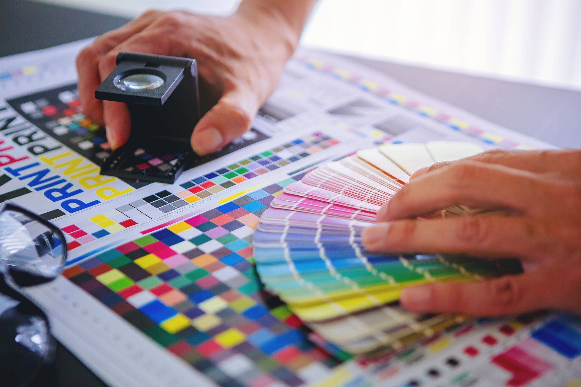 Hands use a magnifying loupe and a color swatch fan to check print quality on a sheet of colorful calibration marks.